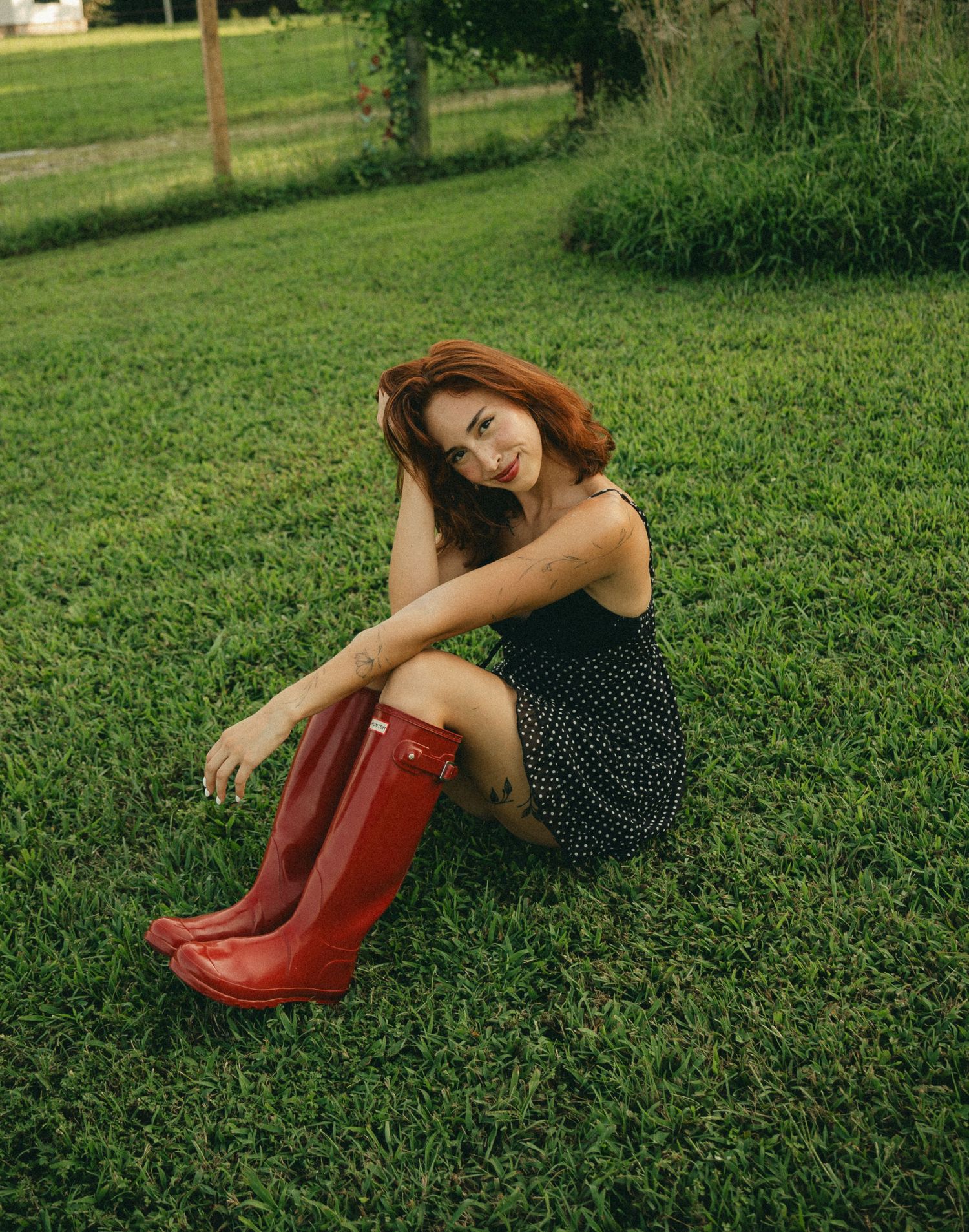 Young woman in polka dot dress and red Hunter rain boots sitting on green grass outdoors.