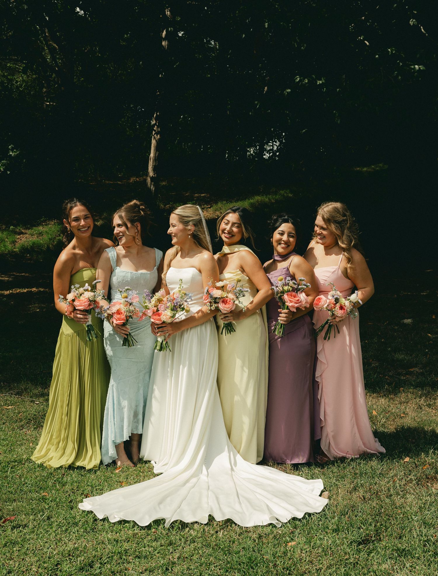 Bride in white gown poses with five bridesmaids in colorful mismatched dresses holding floral bouquets outdoors.