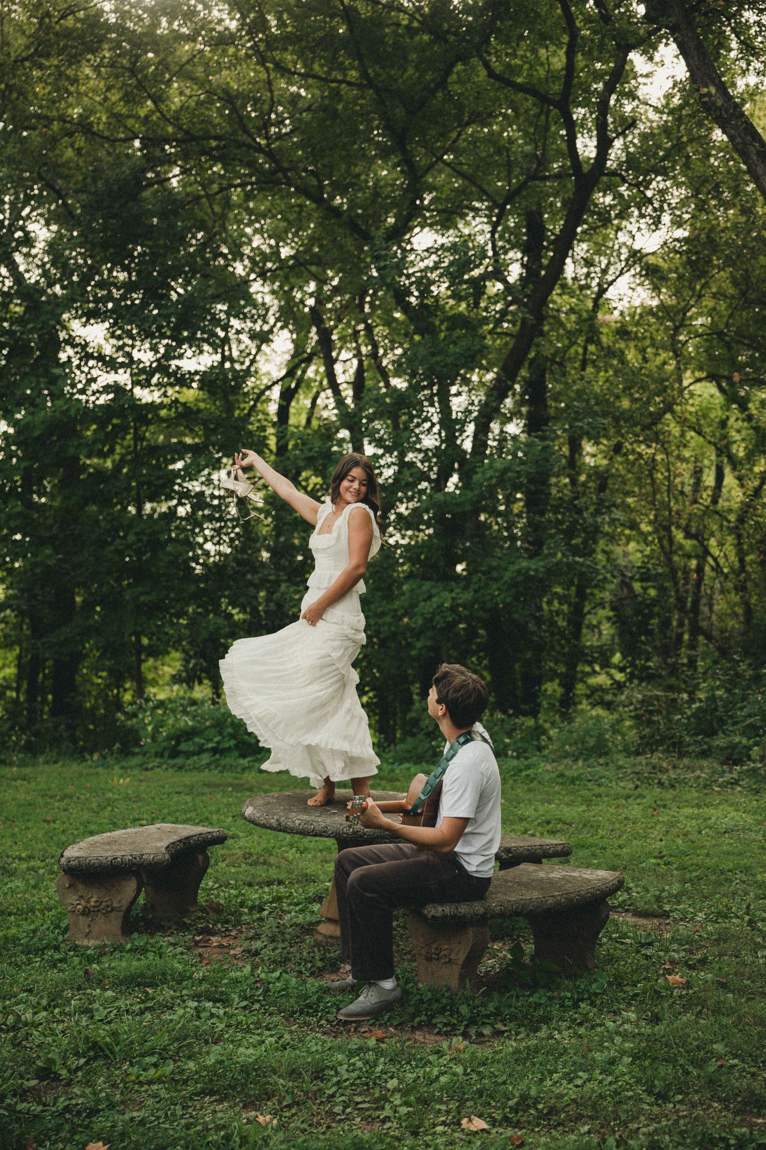 Bride in white dress dancing on stone picnic table while groom sits nearby in lush green park setting.