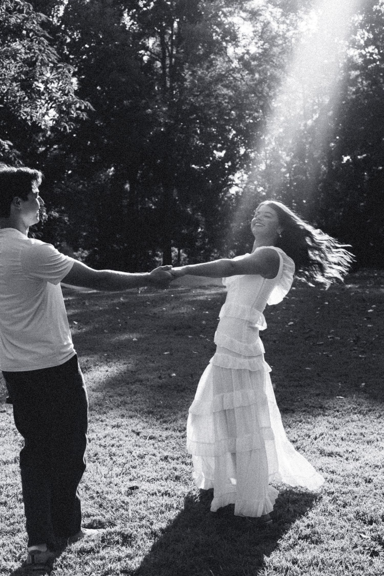 Black and white photo of a couple dancing joyfully in a sunlit park, woman in flowing white dress with hair swirling.
