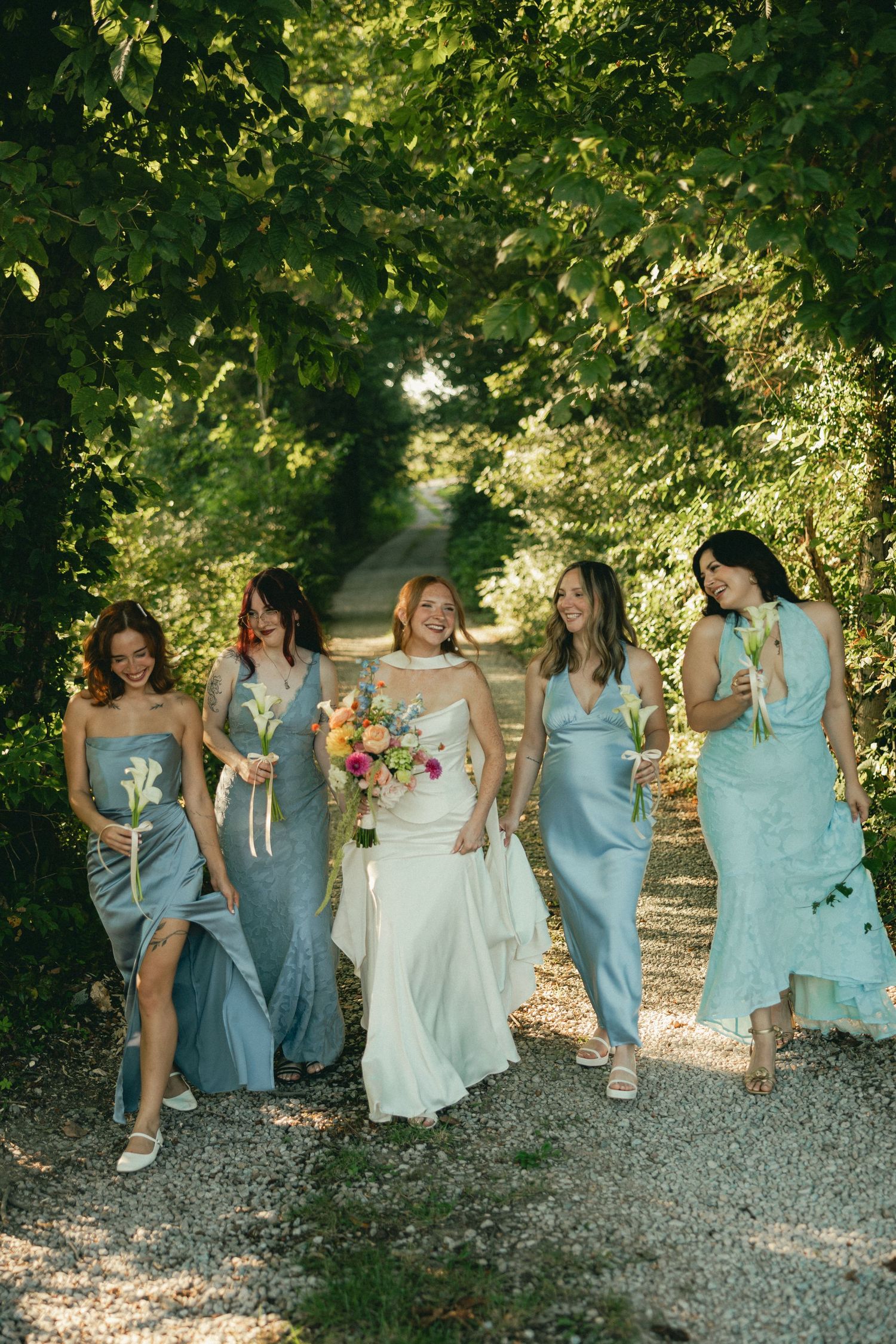 Bride in white gown walks with four bridesmaids in blue dresses along a tree-lined gravel path, holding colorful bouquets.