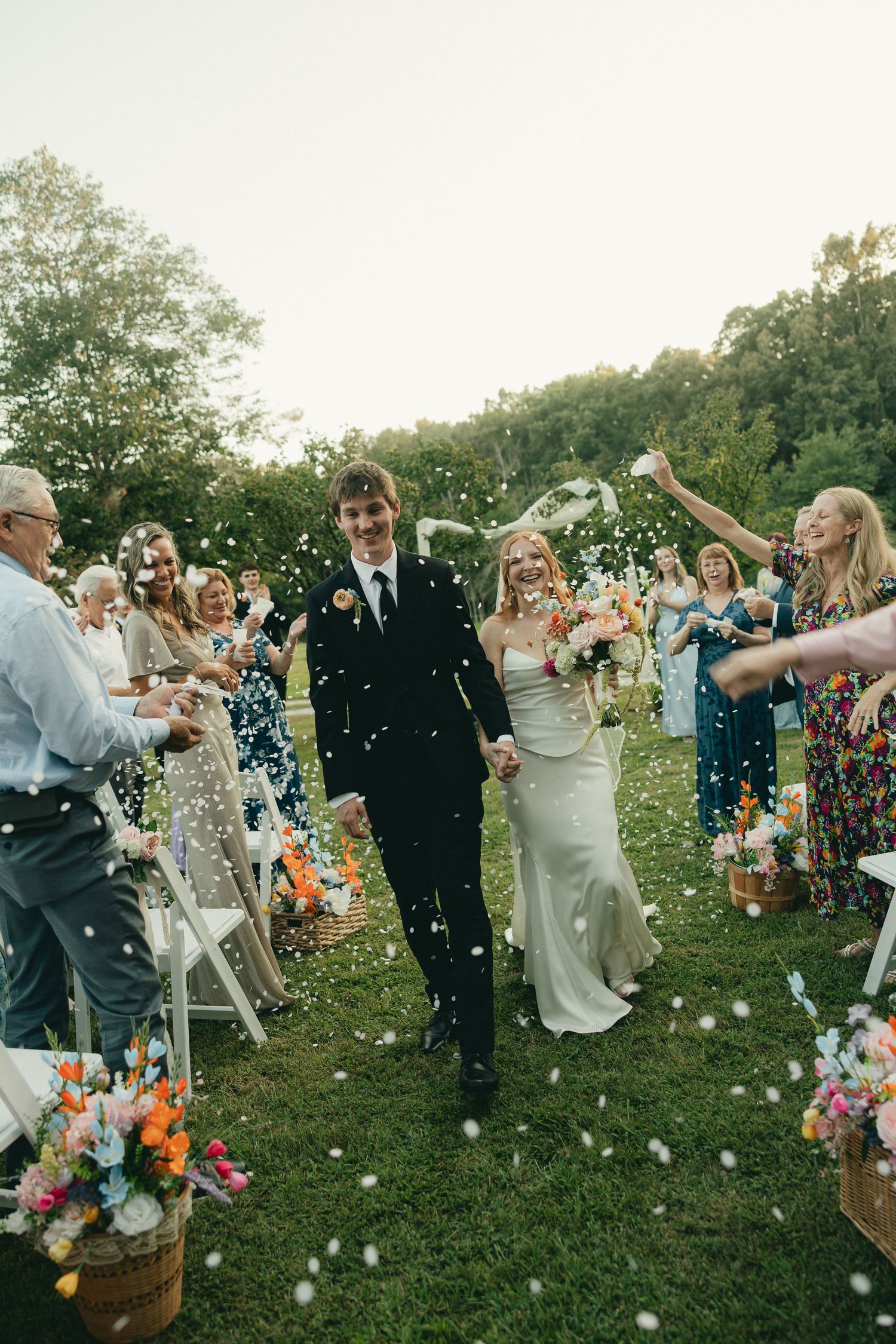 Bride and groom walk down outdoor aisle as guests toss white confetti petals during sunny garden wedding ceremony.