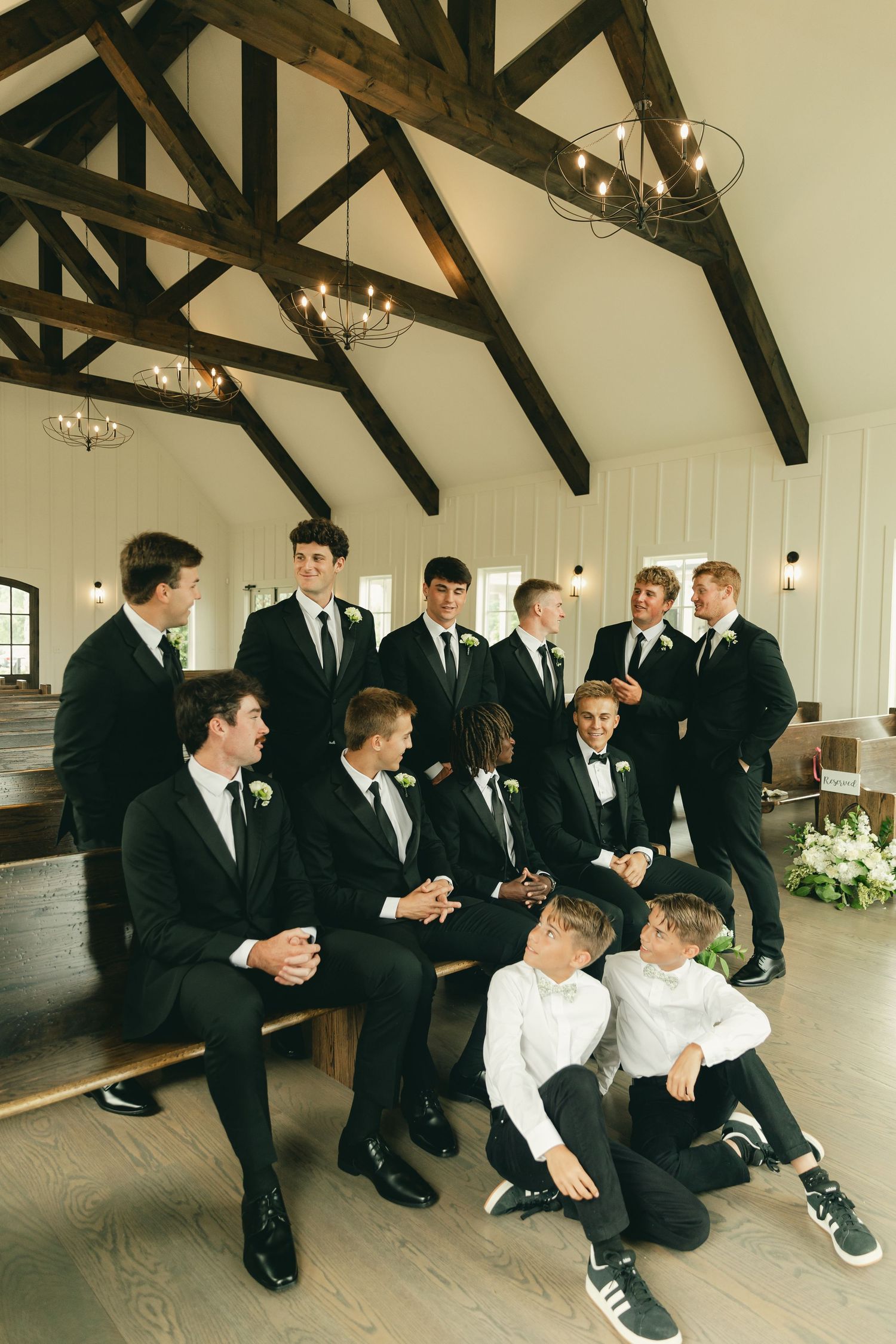 Groom and groomsmen in black suits gather inside a rustic chapel with exposed wood beams.