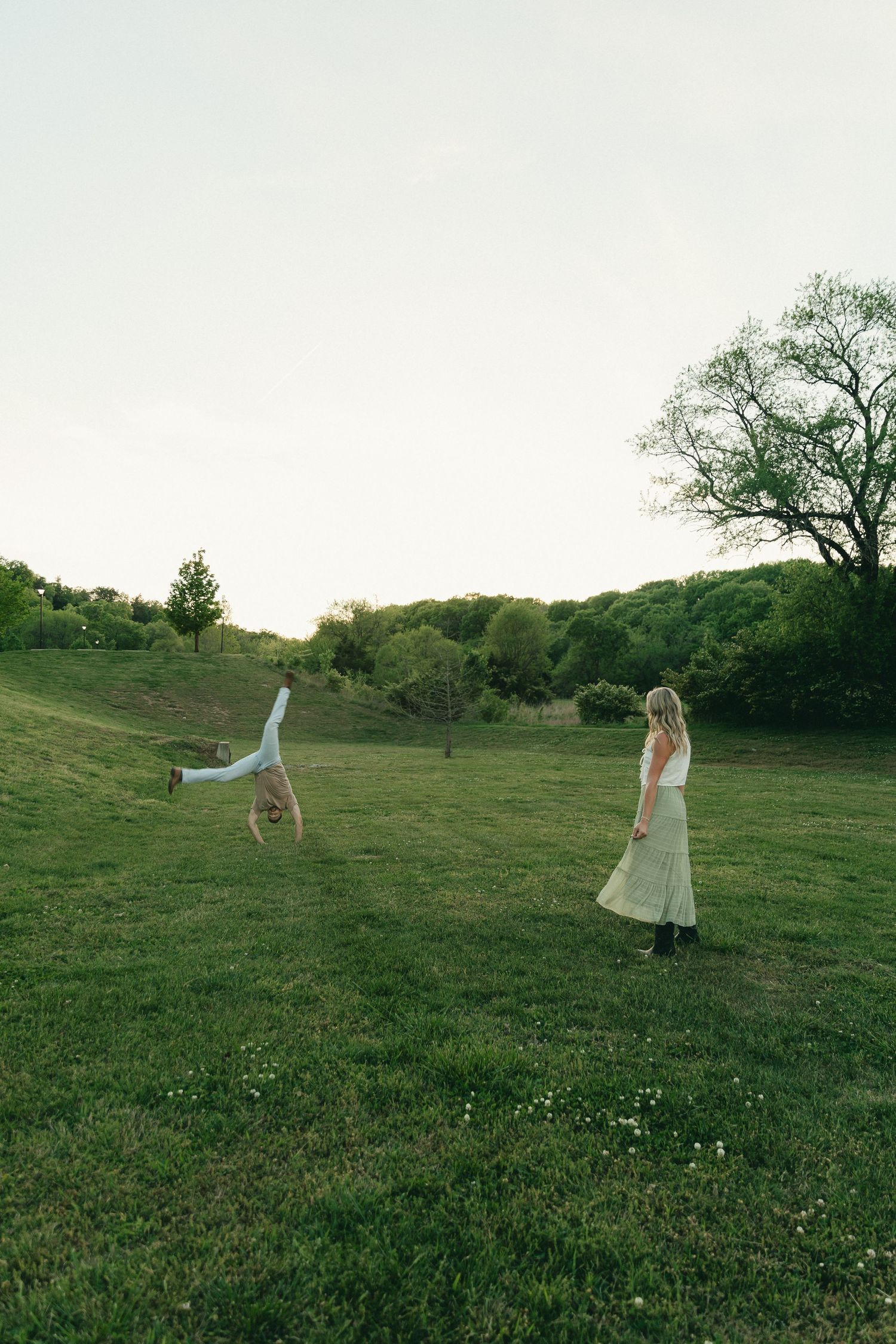 Playful couple in field as man does cartwheel while woman watches on green hillside.