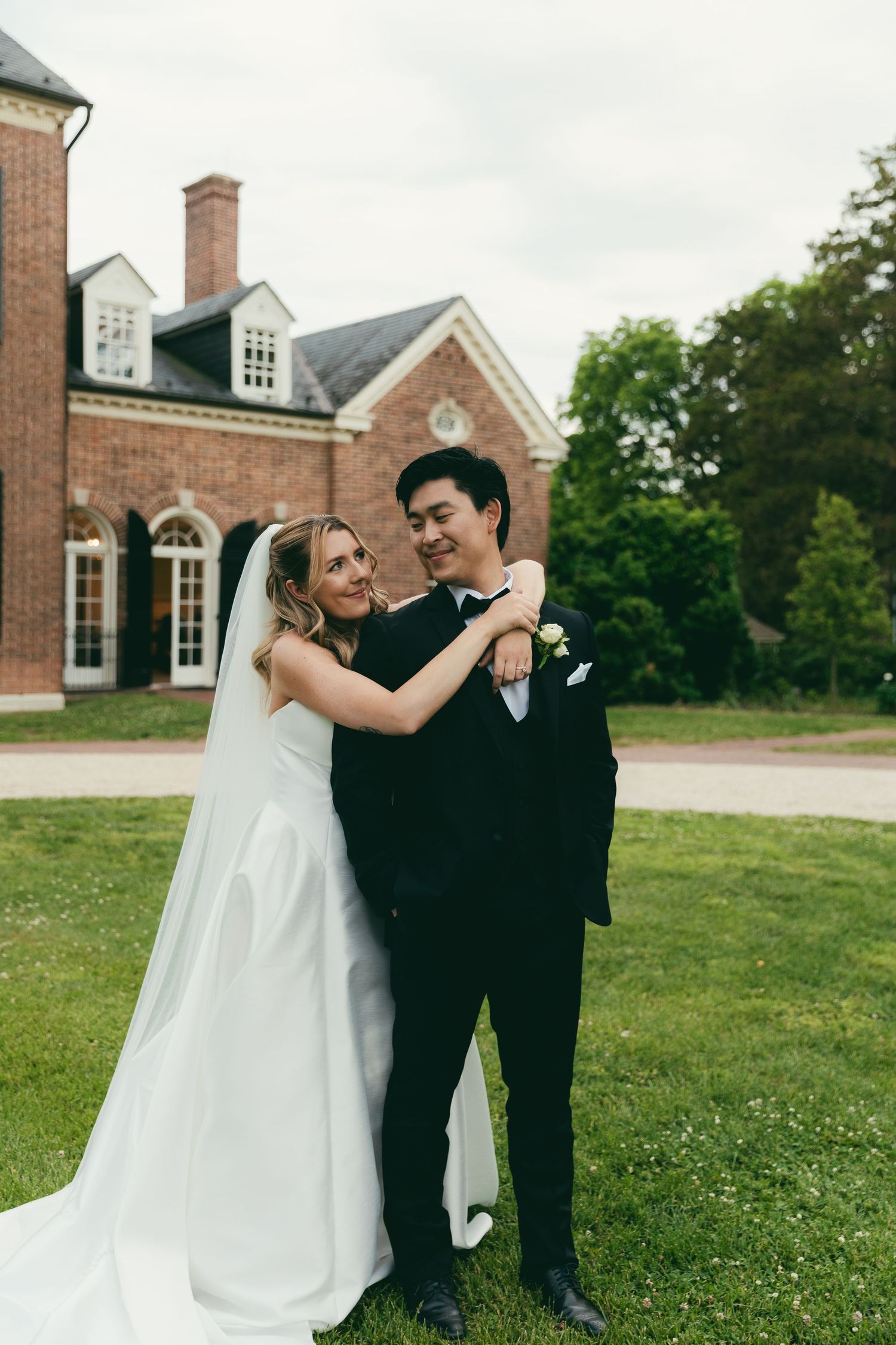Bride in white gown embraces groom in black tuxedo on green lawn before elegant brick manor.