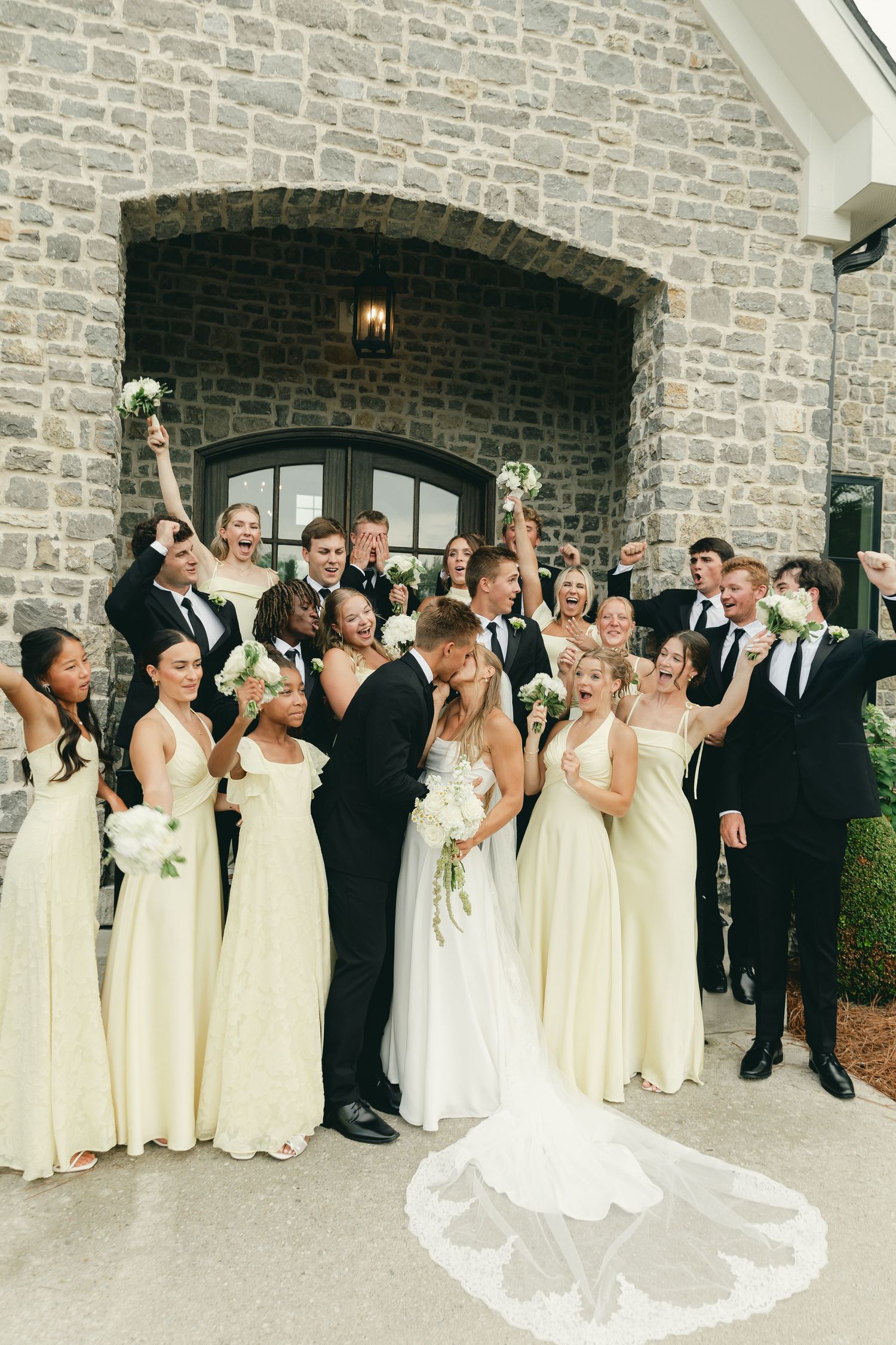 Bride and groom kiss surrounded by cheering wedding party in yellow bridesmaid dresses and black tuxedos outside stone venue.