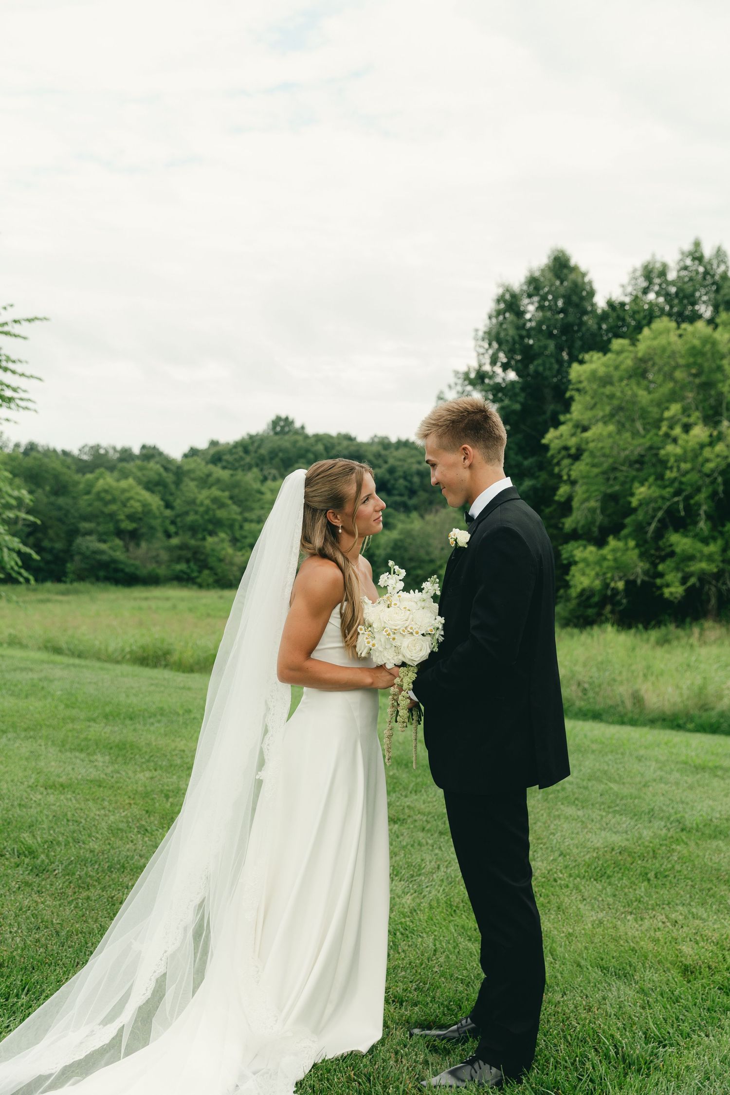 Bride in white gown with cathedral veil and groom in black tuxedo face each other in lush green field.