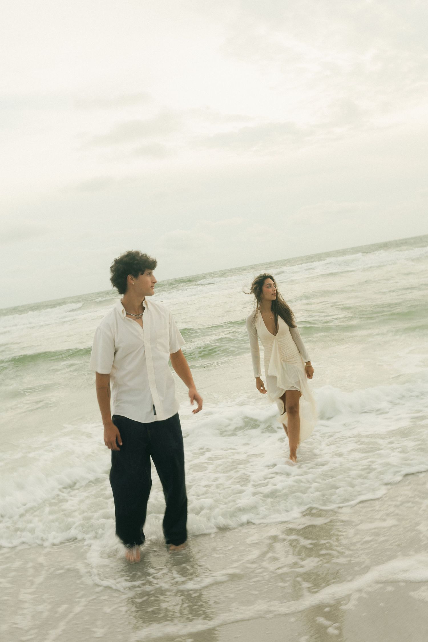 Couple walking through ocean waves on beach at sunset in casual white attire.