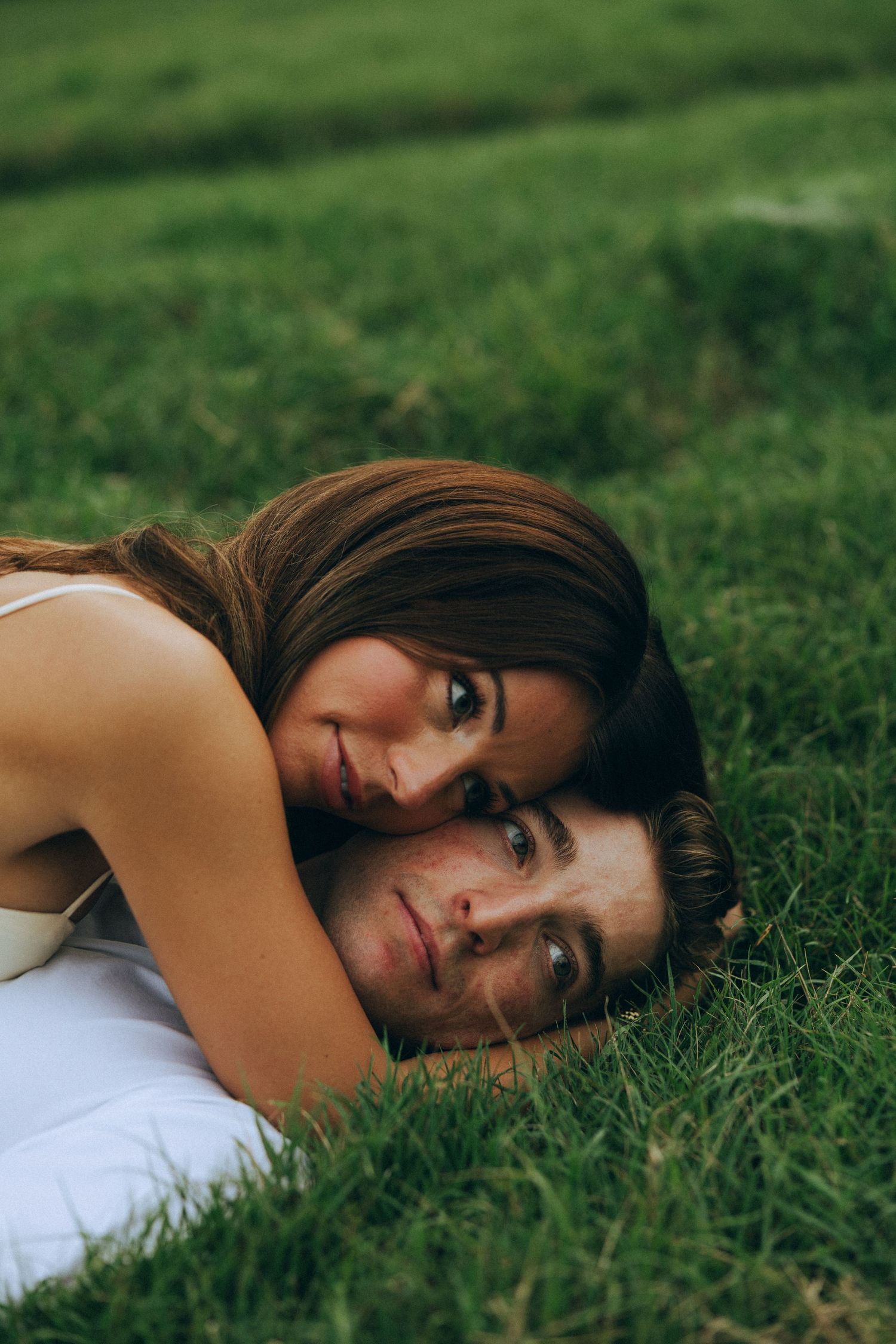 Couple lying on green grass, woman resting her head on man, both looking at camera in a romantic outdoor setting.