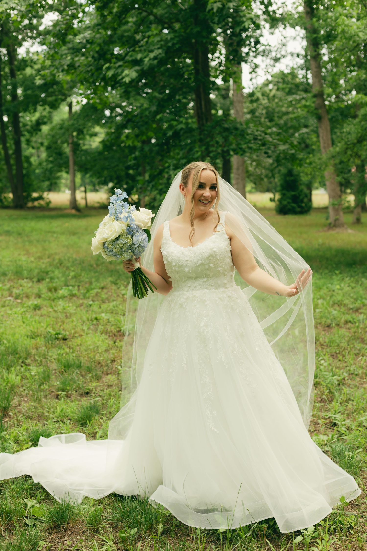 Smiling bride in white lace ball gown holding blue and white bouquet while lifting veil in lush green park.