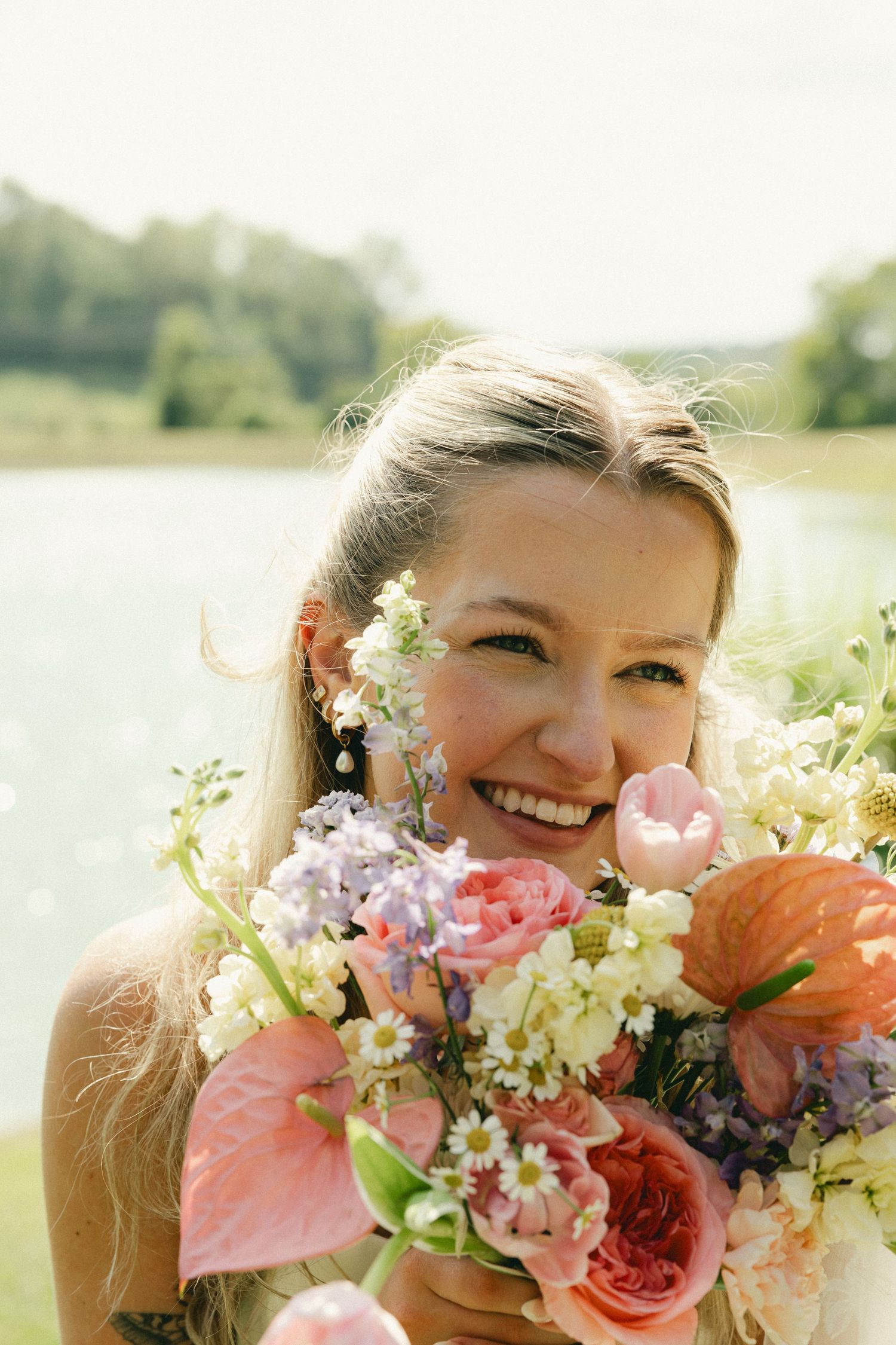 Smiling blonde bride holding a colorful bouquet of pink tulips, roses, and wildflowers near a serene lake.