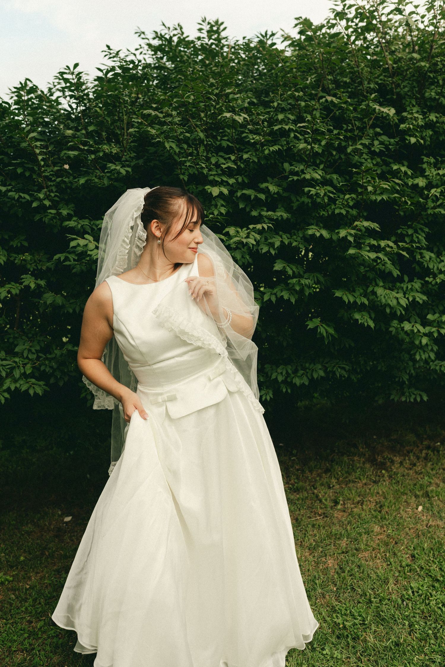 Elegant bride in white sleeveless gown and veil posing in front of lush green hedge.