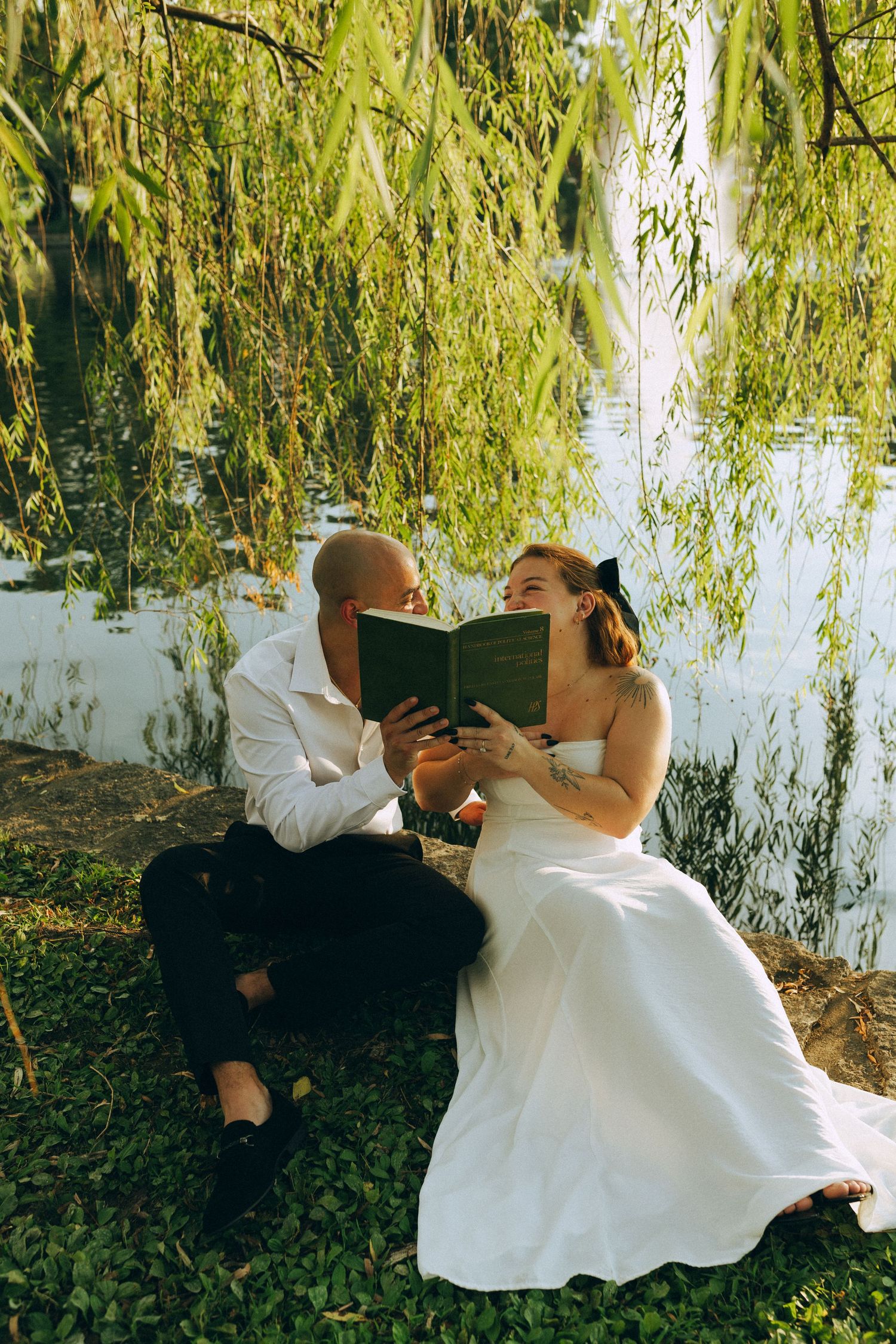 A bride and groom sit under a weeping willow tree by a lake, sharing a romantic kiss behind an open book.