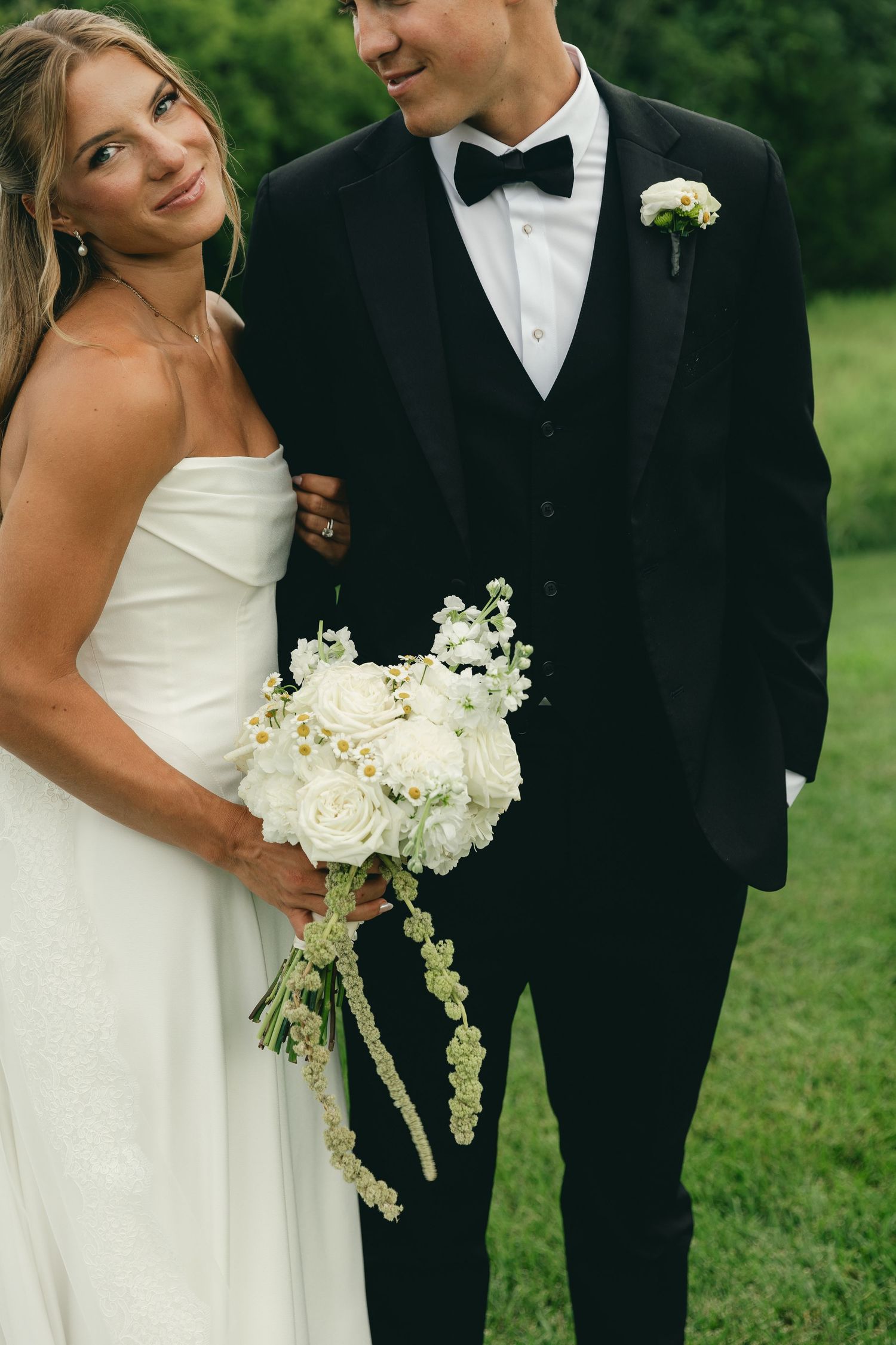 Bride in strapless white gown holds bouquet of white roses beside groom in black tuxedo outdoors.