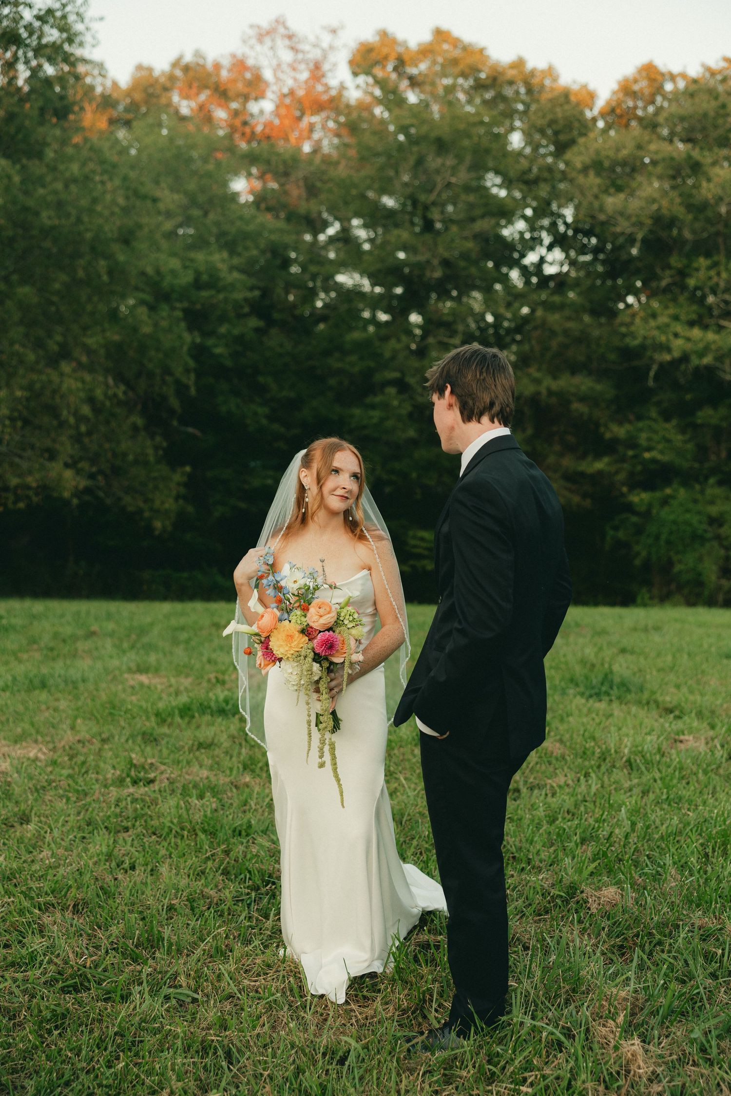 Bride in white gown holding colorful bouquet faces groom in black suit on green lawn with autumn trees backdrop.