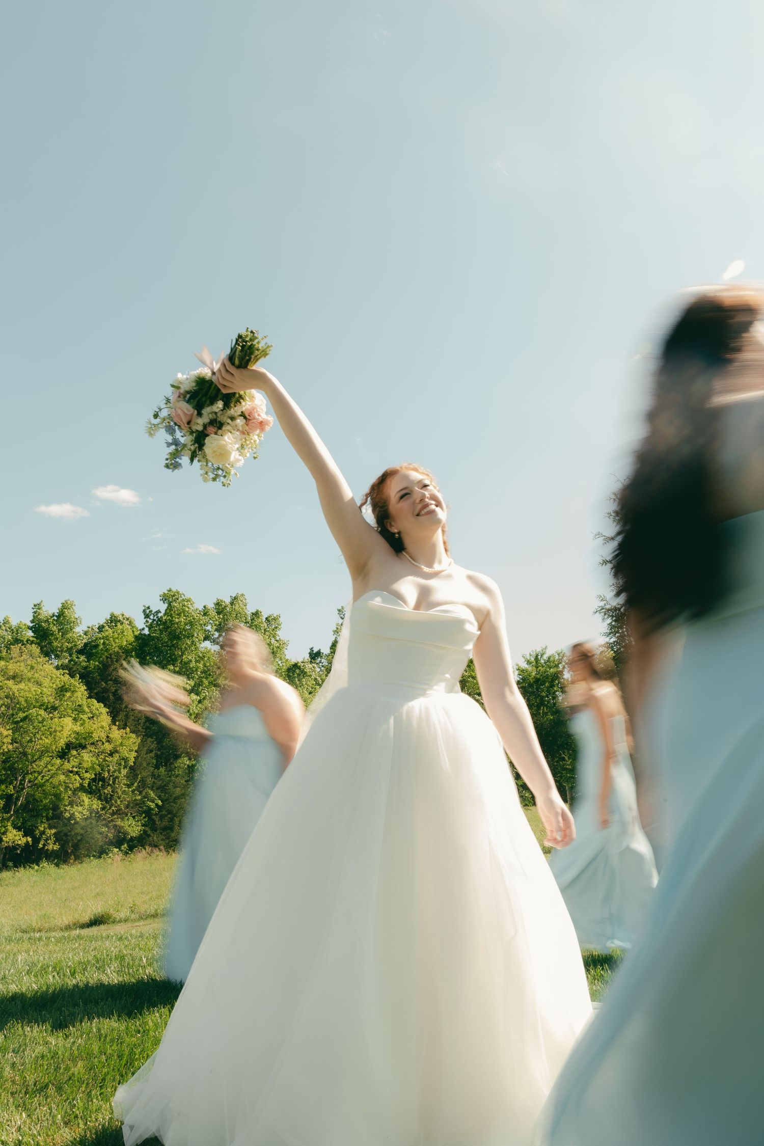 Radiant bride in white gown raising bouquet overhead while bridesmaids in blue dresses celebrate outdoors.