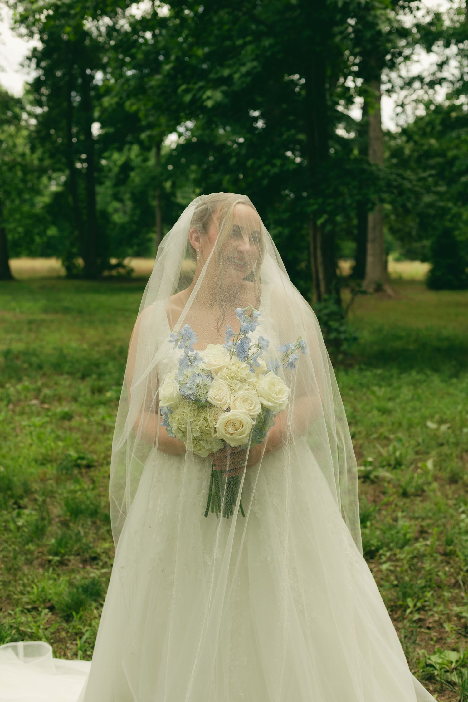 Bride smiling under sheer veil holding white roses and blue hydrangea bouquet in green park.