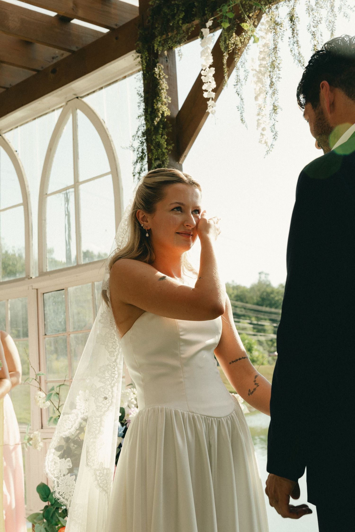 Emotional bride wiping tears in white gown and veil, holding groom's hand during wedding ceremony in greenhouse venue.