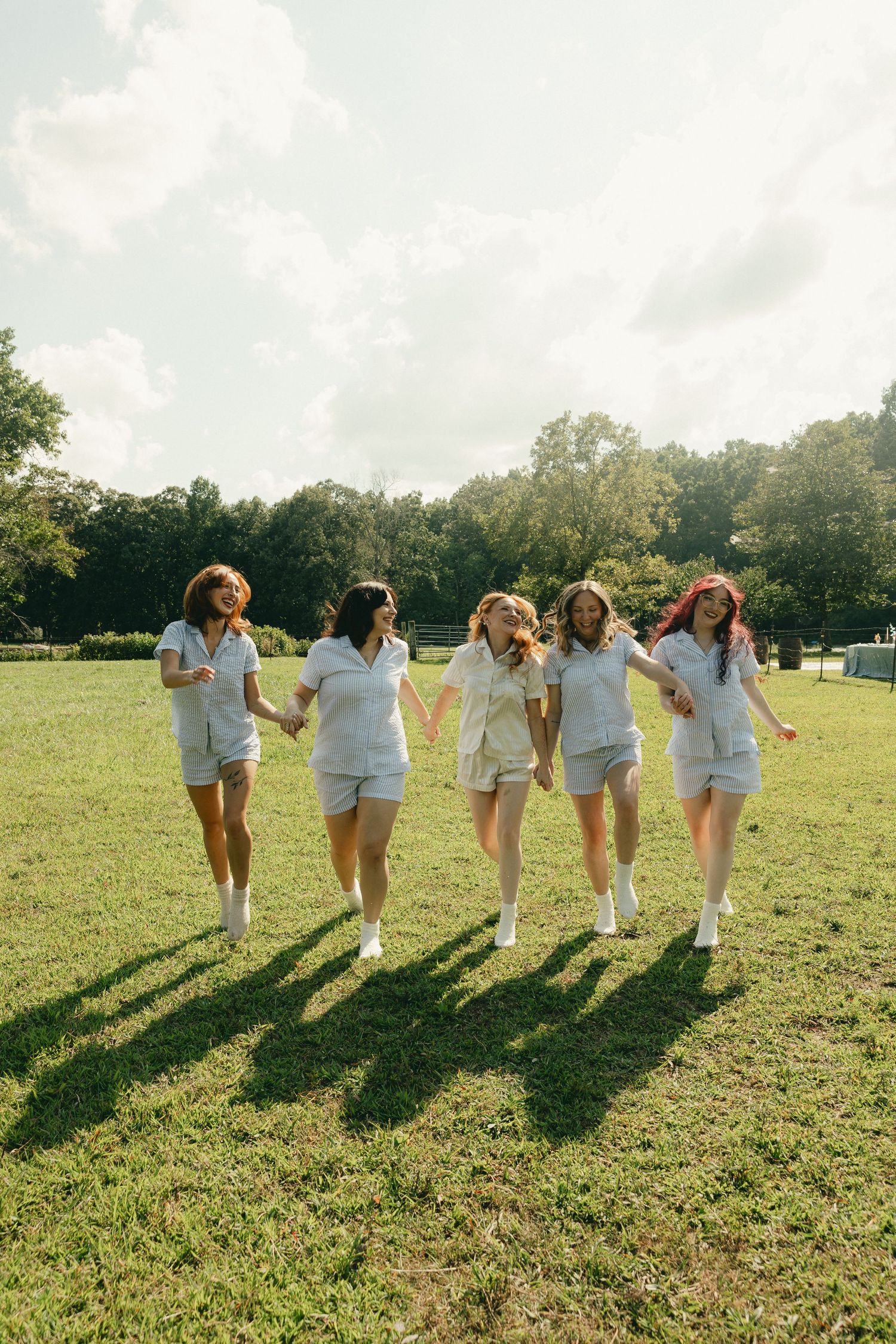 Five young women in matching white outfits hold hands while walking across a sunny green field.