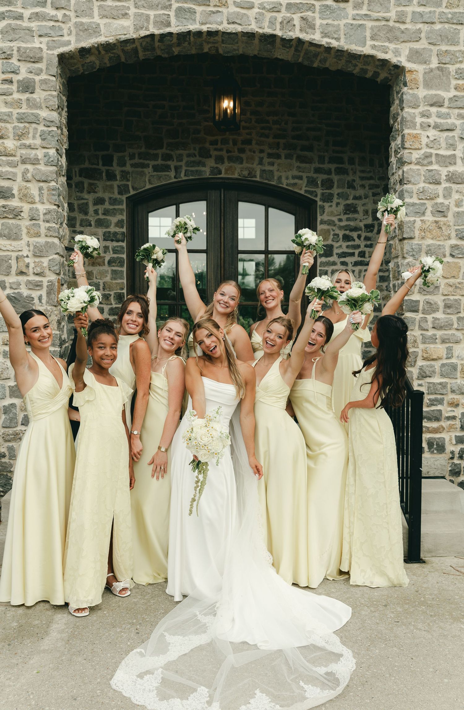 Bride poses with bridesmaids in light yellow gowns raising bouquets in celebration outside a stone building.