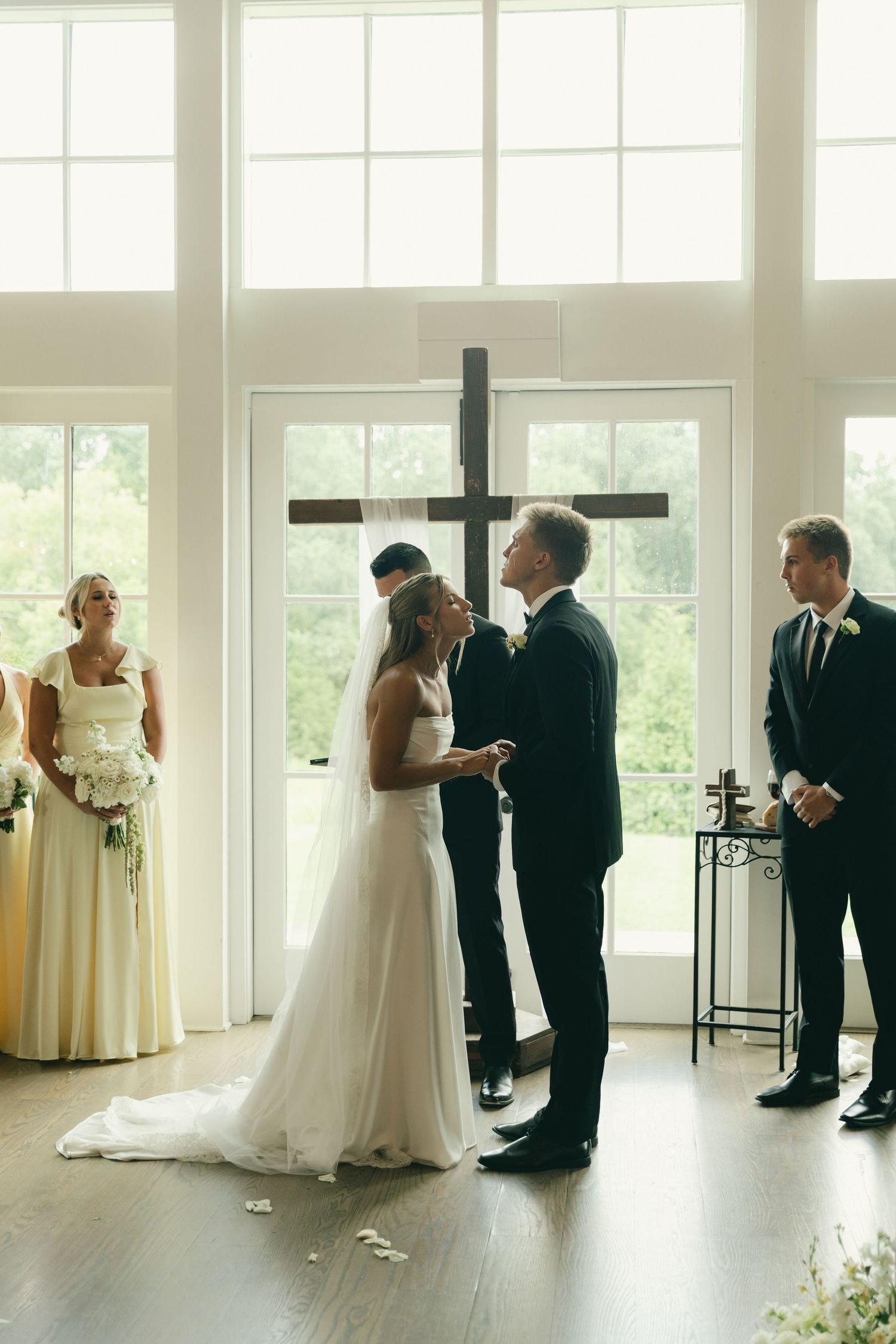 Bride and groom share first kiss at altar before large wooden cross in sunlit white wedding chapel.