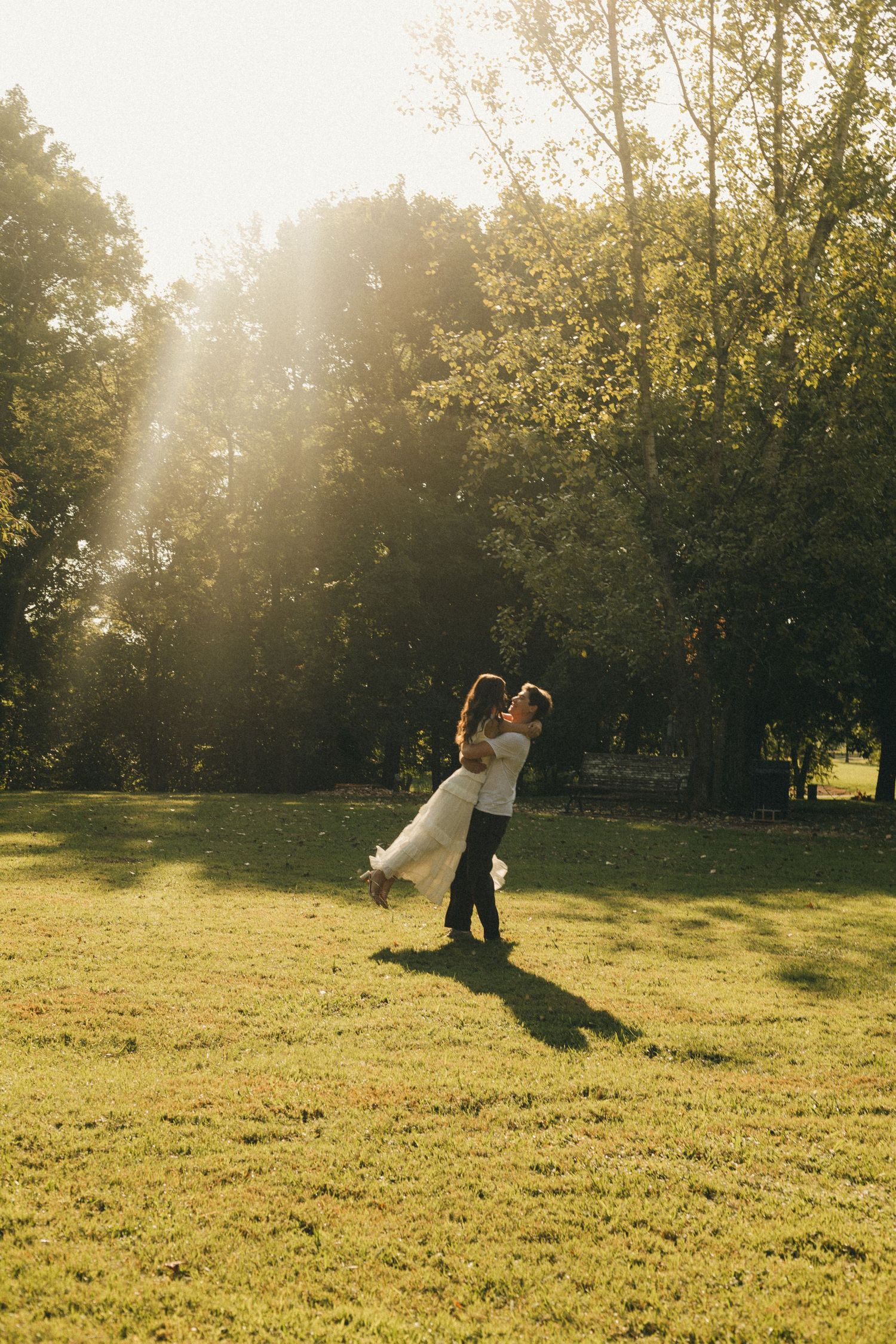 Groom lifting bride in sunlit park, golden hour light filtering through trees on lush green grass.