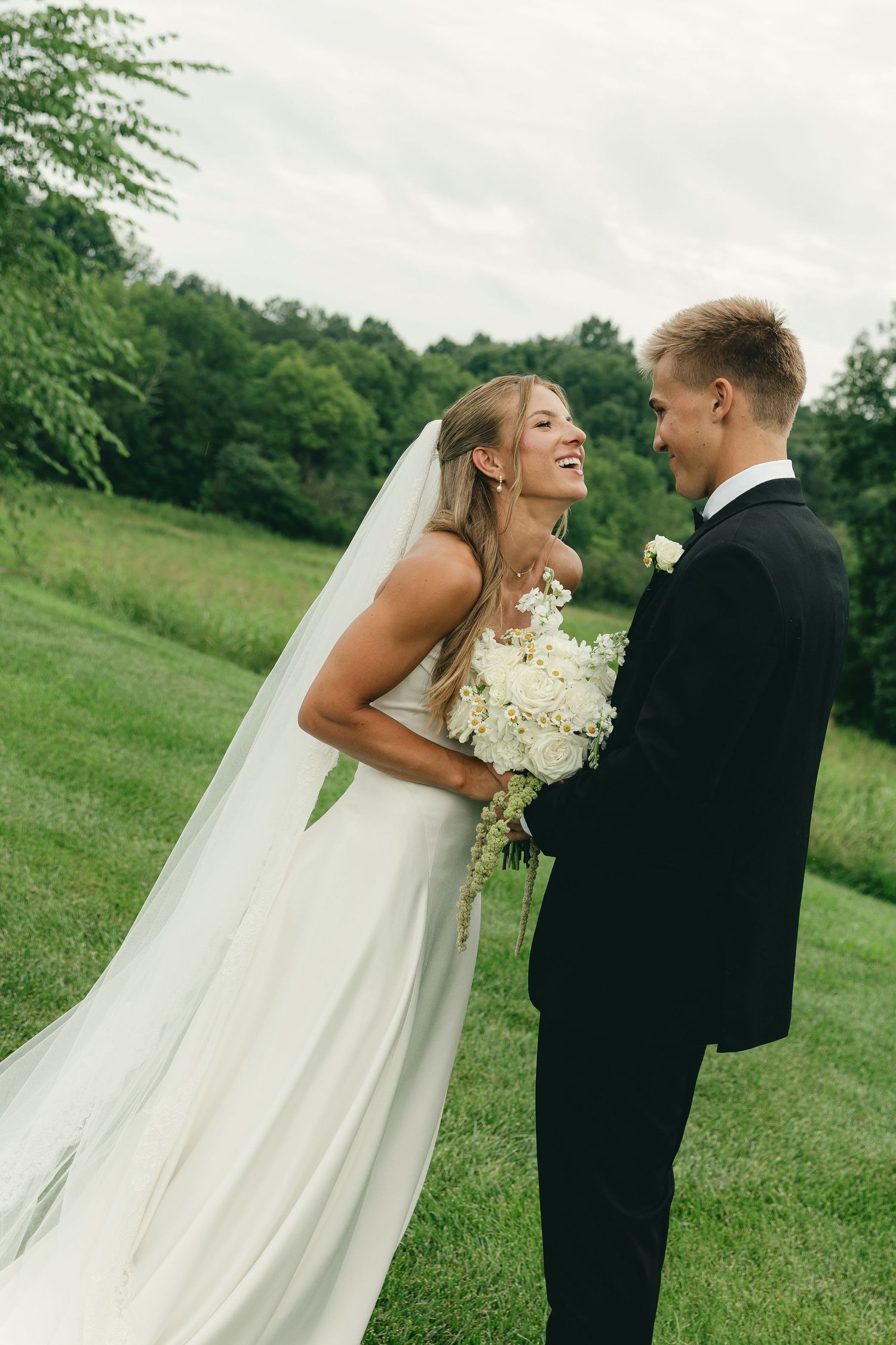 A smiling bride in a white gown and veil holds a bouquet while facing her groom in a black tuxedo outdoors.