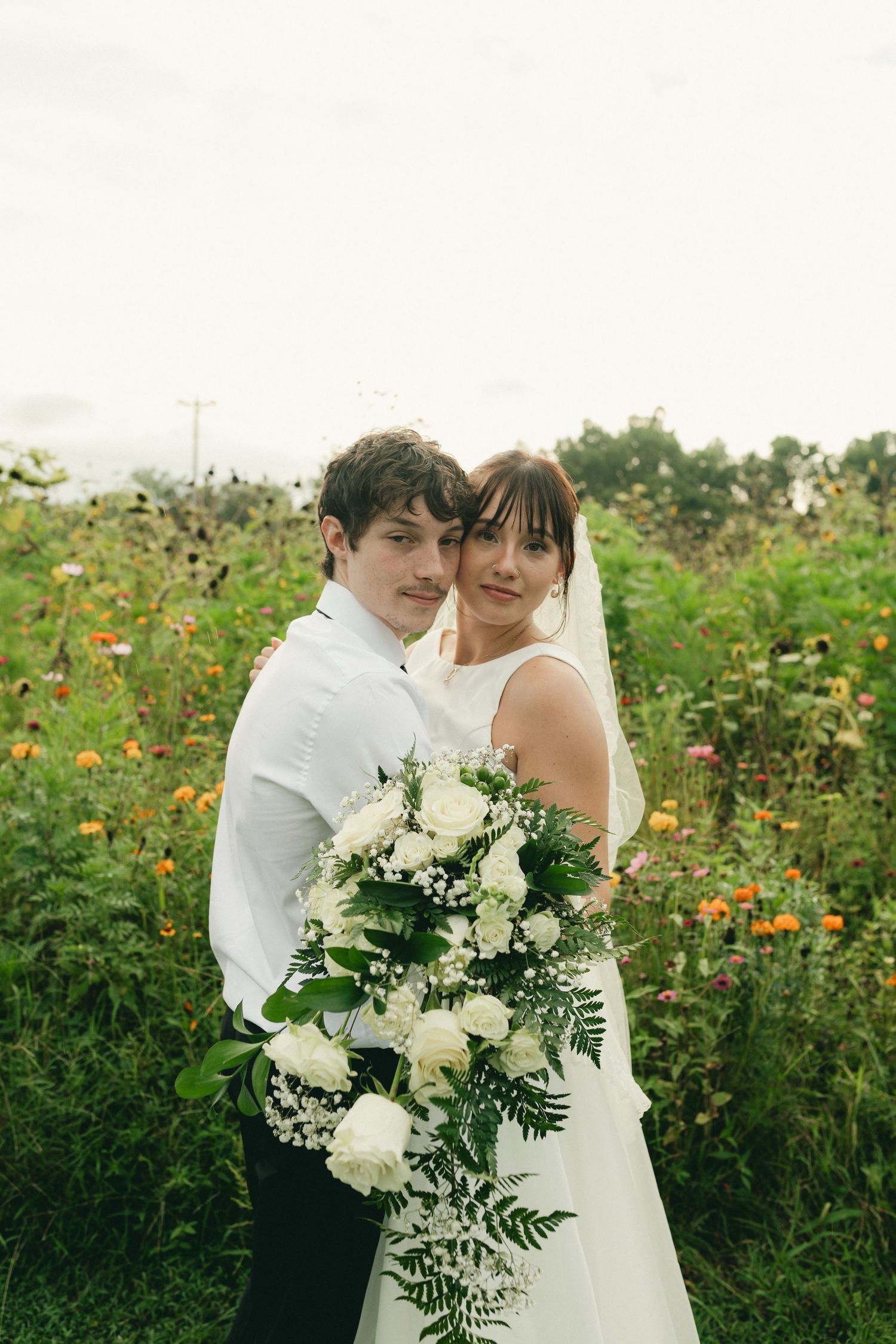 Bride and groom embracing in wildflower field holding white rose bouquet on wedding day.