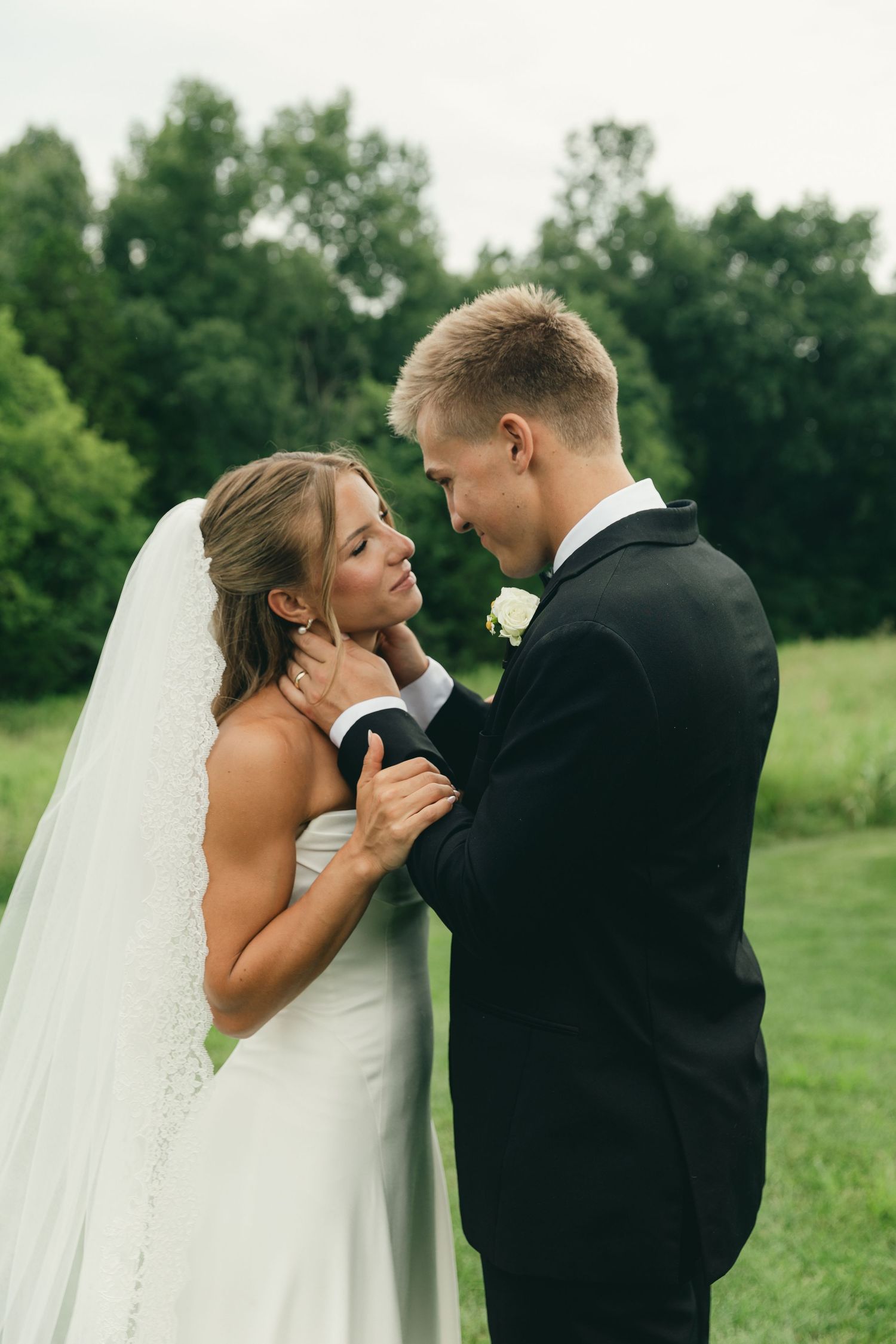 Bride and groom share an intimate moment, groom tenderly holding bride's face outdoors with lush green trees behind them.