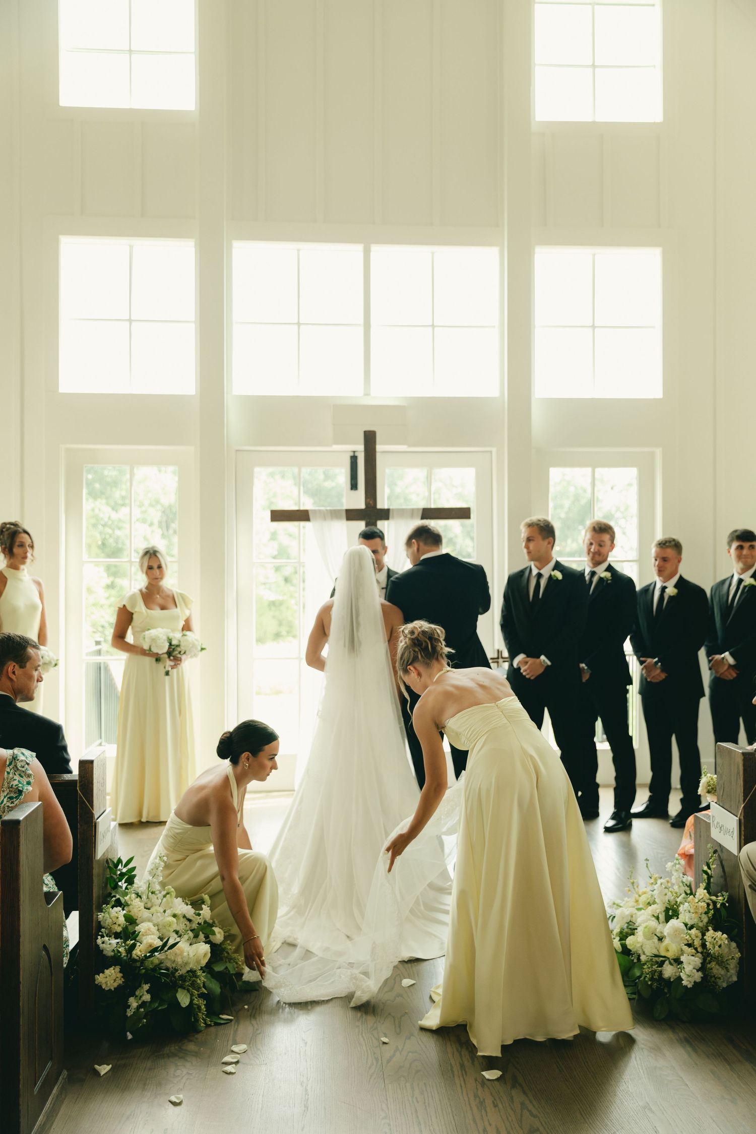 Bridesmaids arrange bride's cathedral veil and train at altar during wedding ceremony in white chapel.