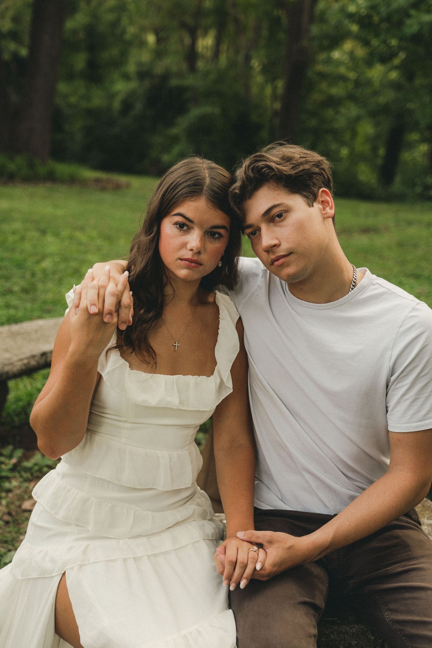 Young couple in white outfits sitting closely together on bench in green park, holding hands tenderly.