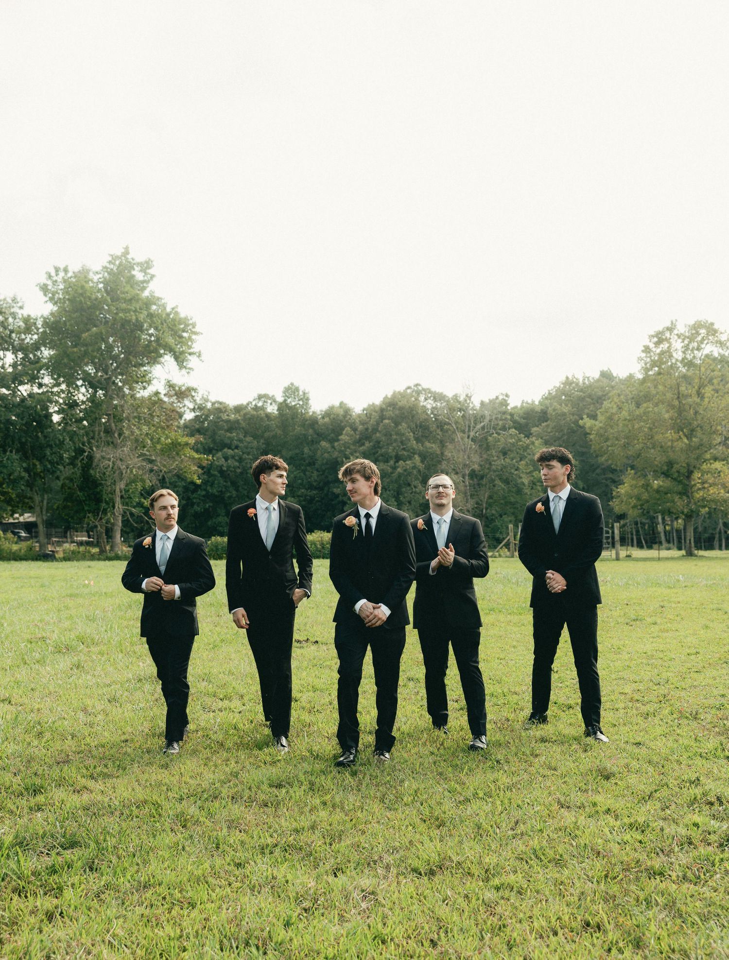 Five young men in black suits and bow ties pose in a green field with trees in the background at a wedding.