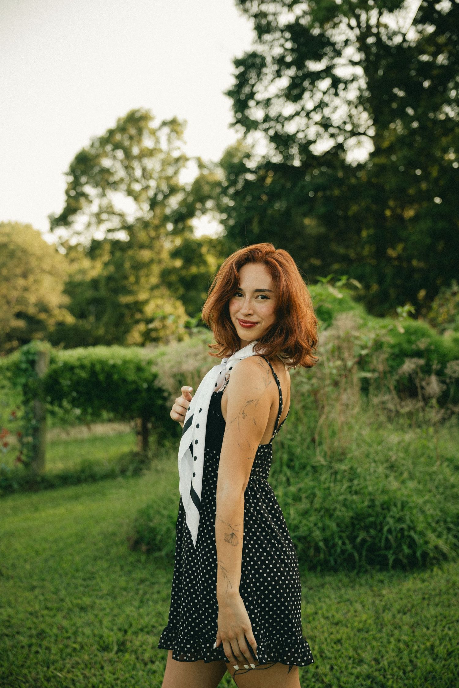 Smiling redhead woman in polka dot dress with white scarf posing in a lush green garden.