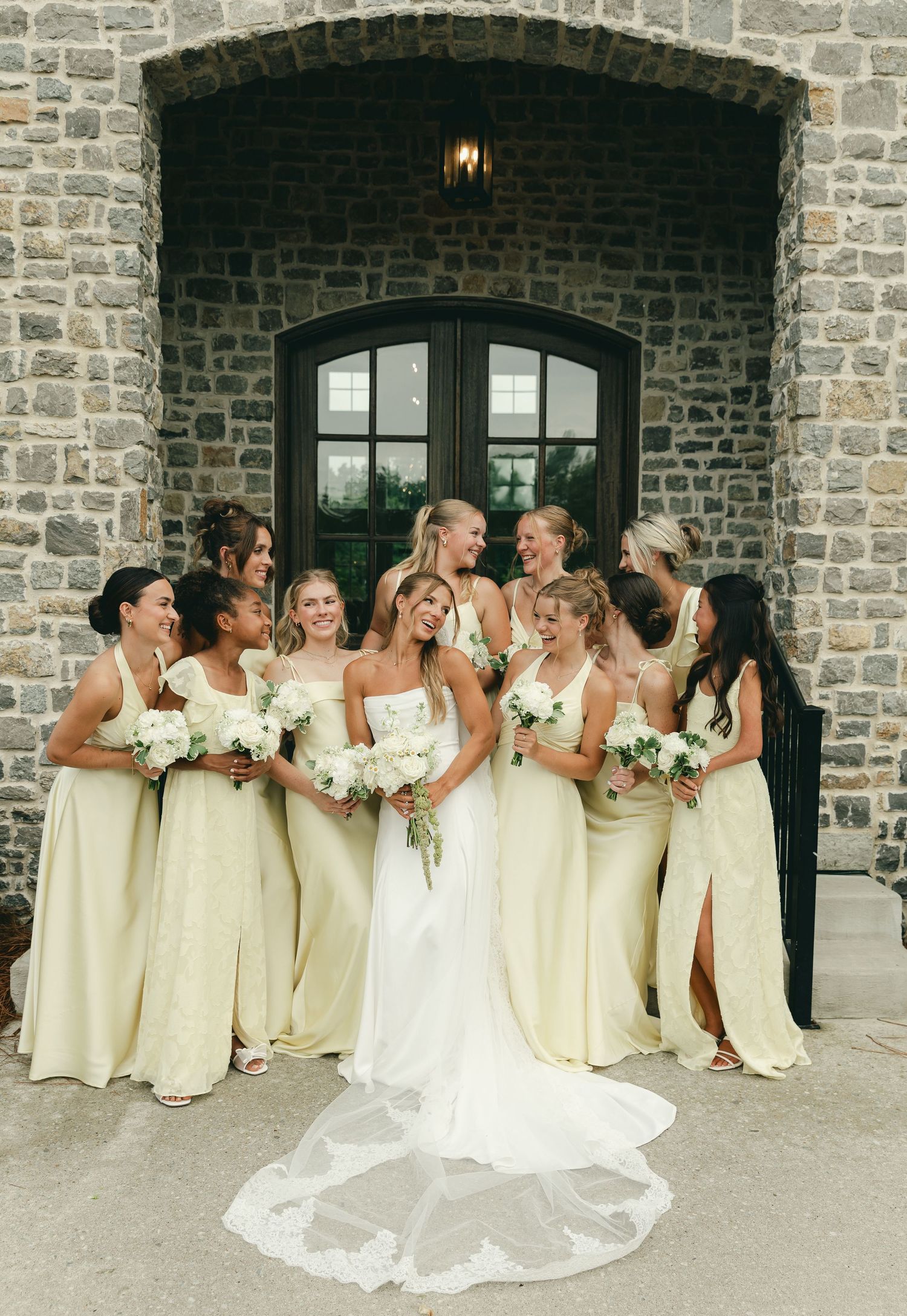 Bride in white gown with train poses with bridesmaids in yellow dresses outside stone building.