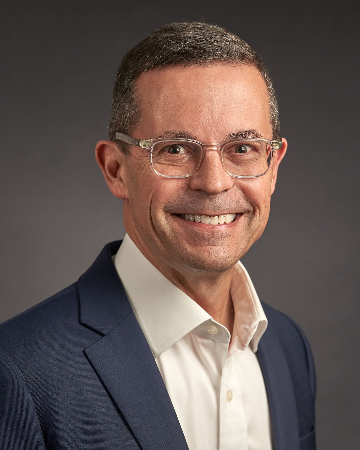Executive business portrait of a man in navy suit and white shirt against dark background.
