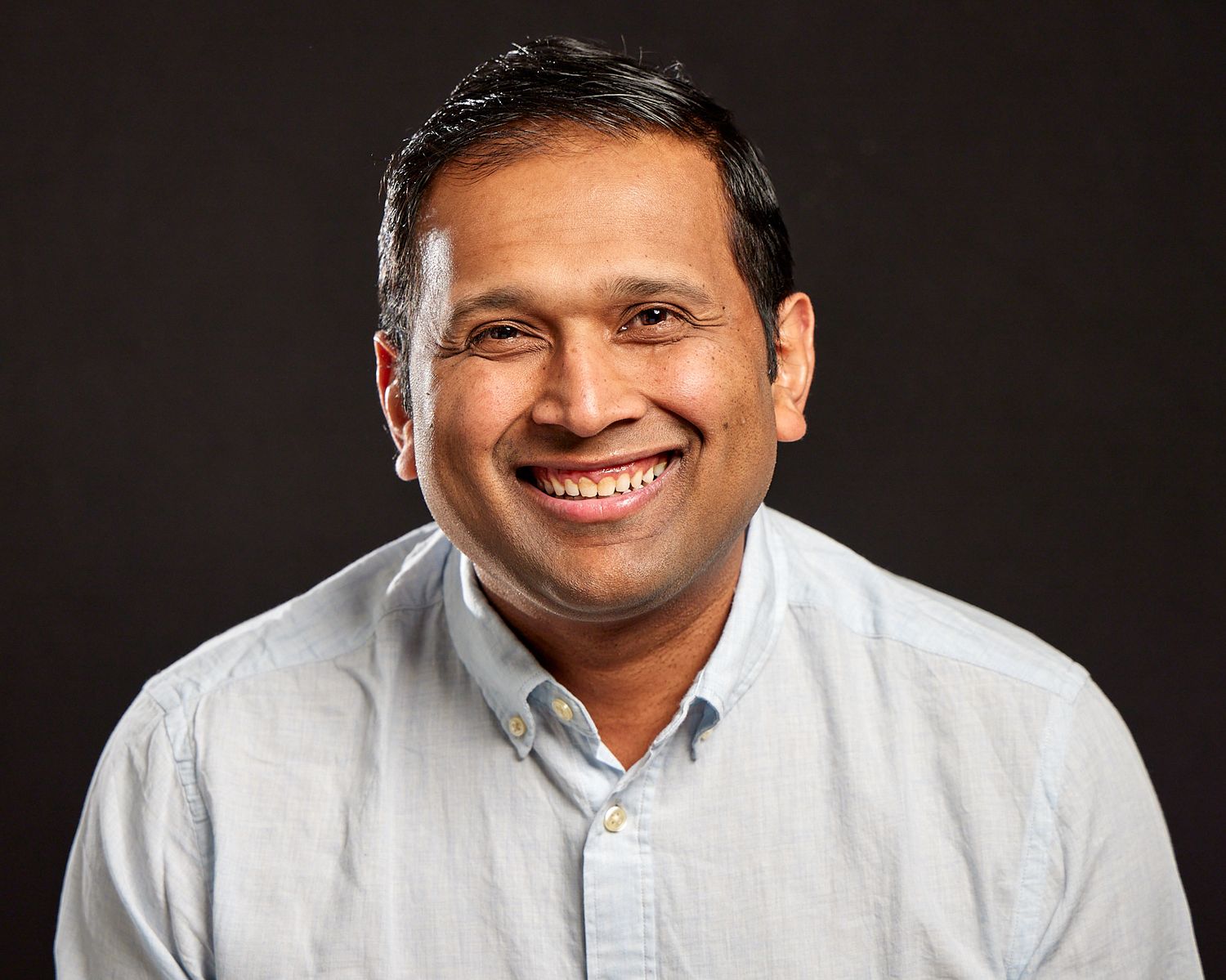 Professional headshot of someone in a light gray button-down shirt against a dark background.