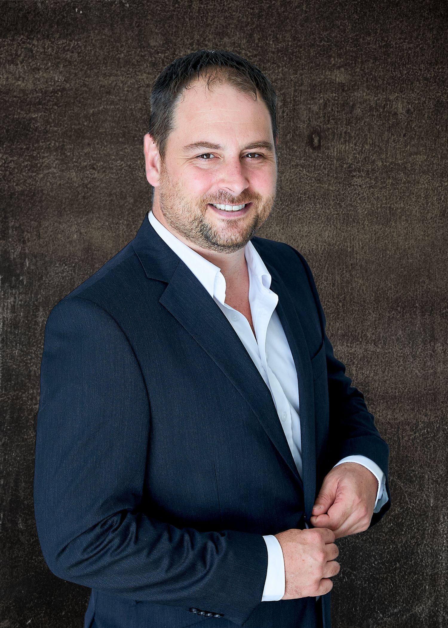 A headshot photograph - Professional man in dark suit showing casual pose against textured background.