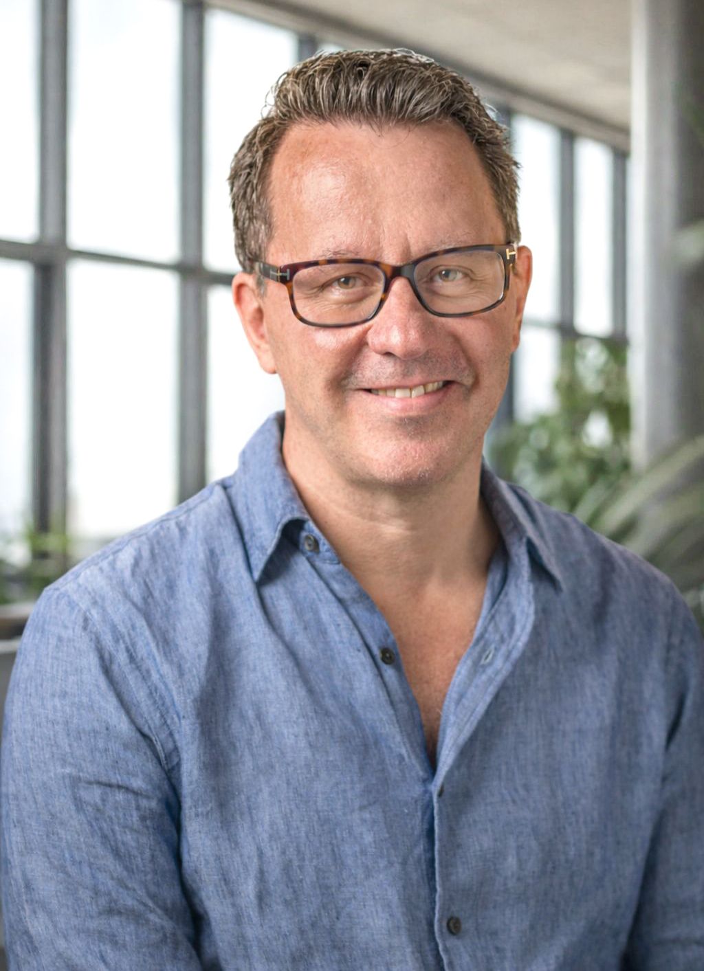 Casual business portrait of someone in denim shirt against window-lit office background.