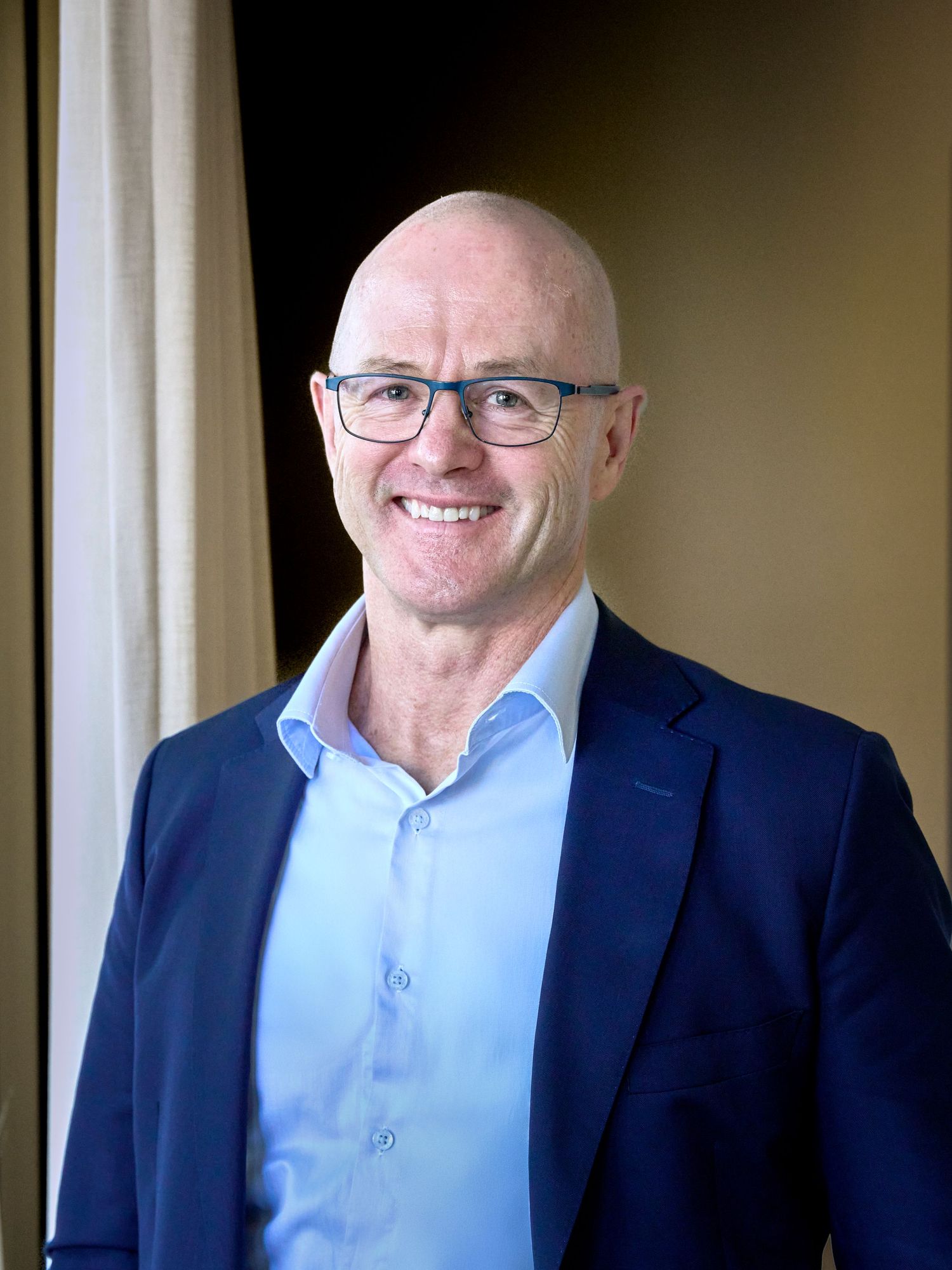 Professional portrait of a man in glasses and blue suit standing by a window.