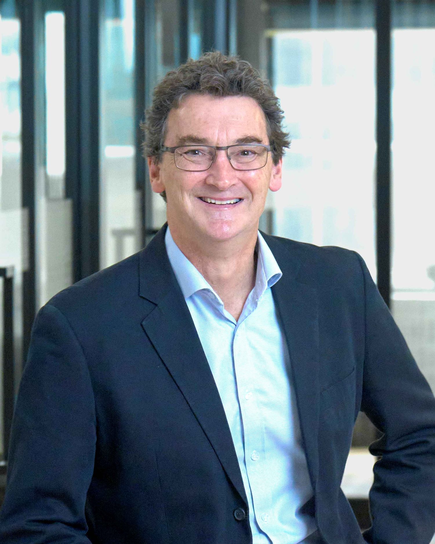 Professional business headshot of someone in navy blazer and blue shirt in office setting.
