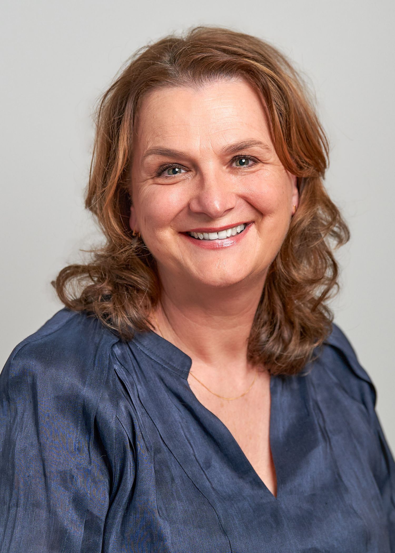 Professional headshot of someone with wavy brown hair wearing a navy blue blouse against a light gray background.