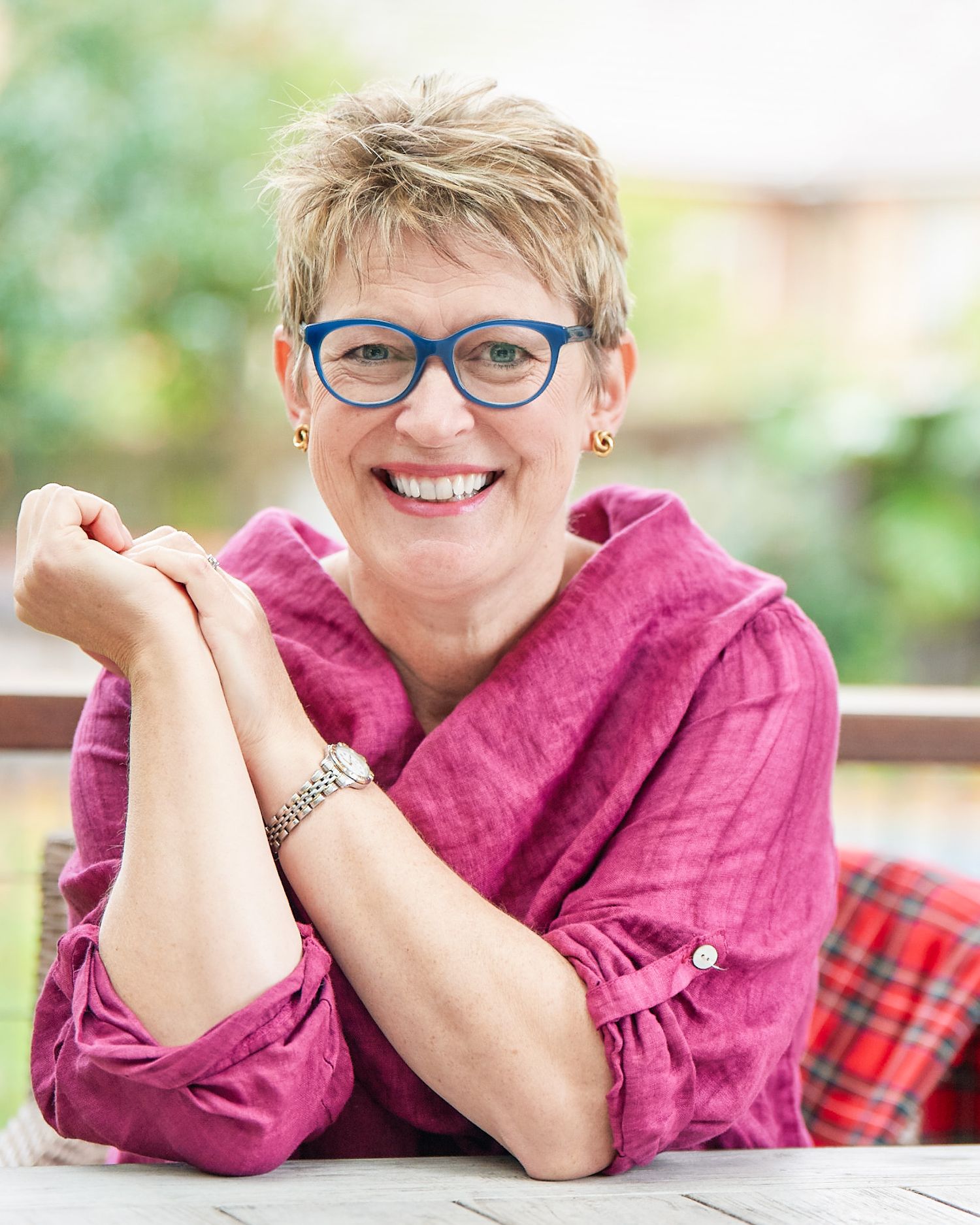 Headshot of of person in pink sweater and glasses smiling while resting head on hands in outdoor setting.
