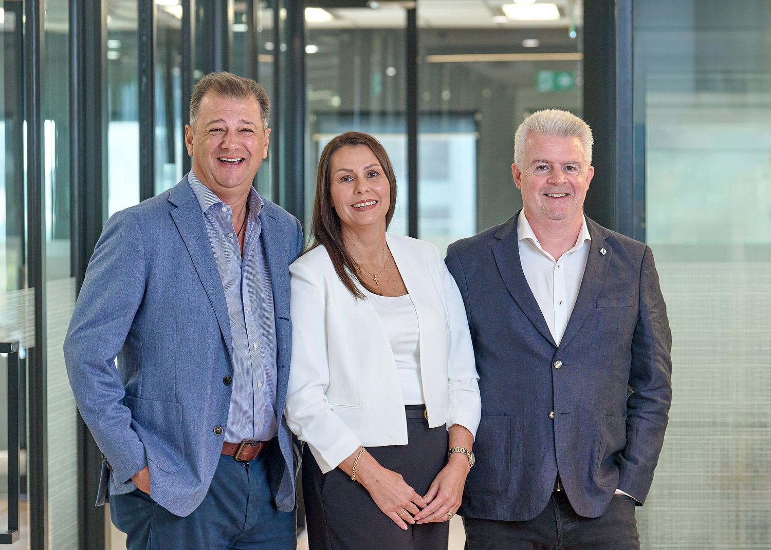Professional business team in suits and business attire standing together in modern office hallway with glass walls.