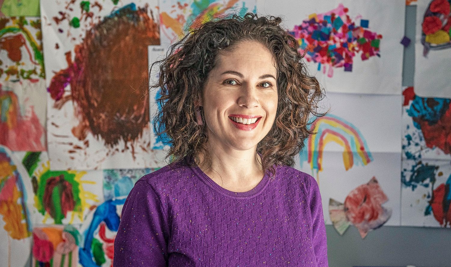 Causal professional portrait of someone in a purple sweater standing in front of colorful children's artwork.