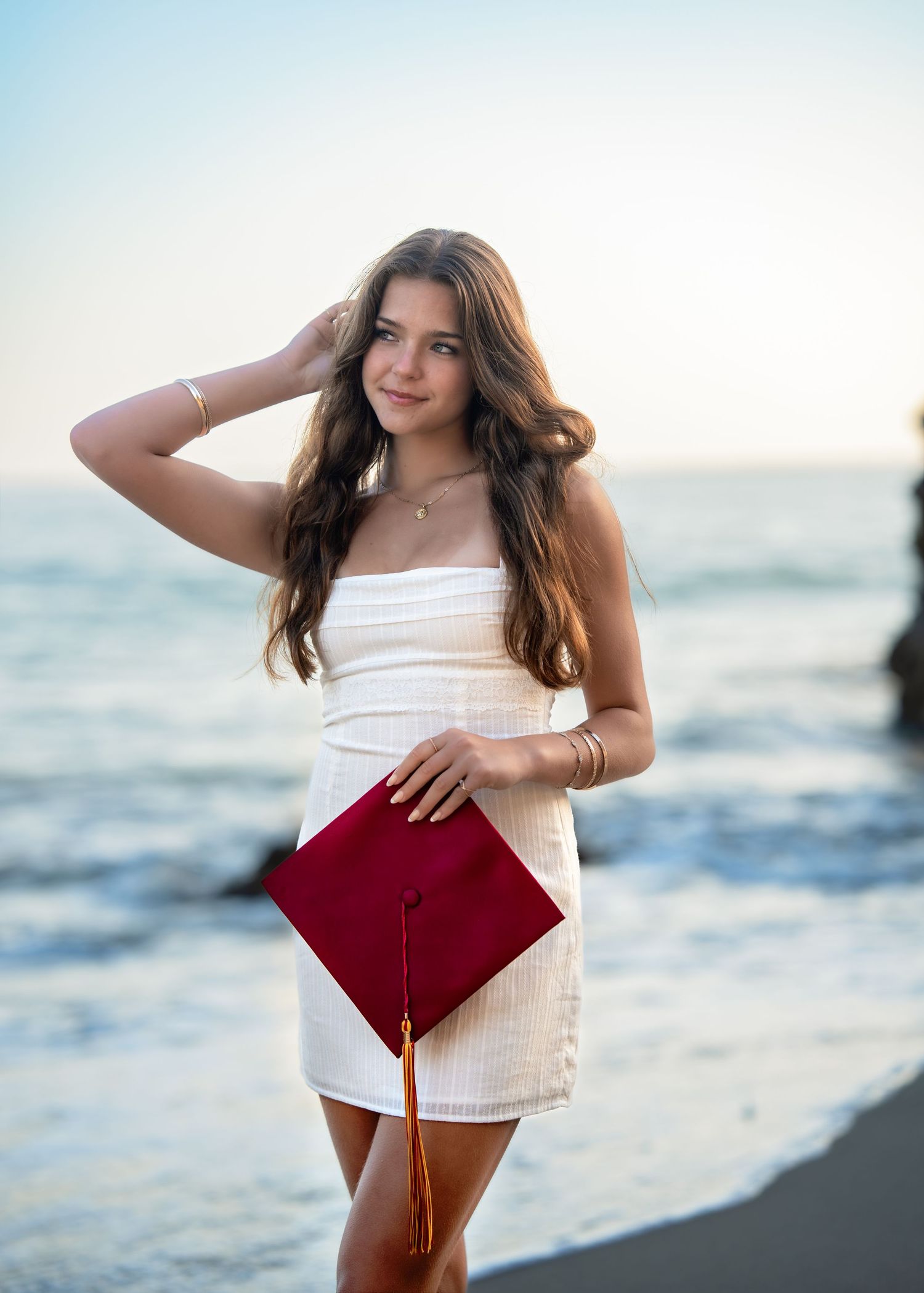 Young woman in white dress holding red graduation cap on beach at sunset.