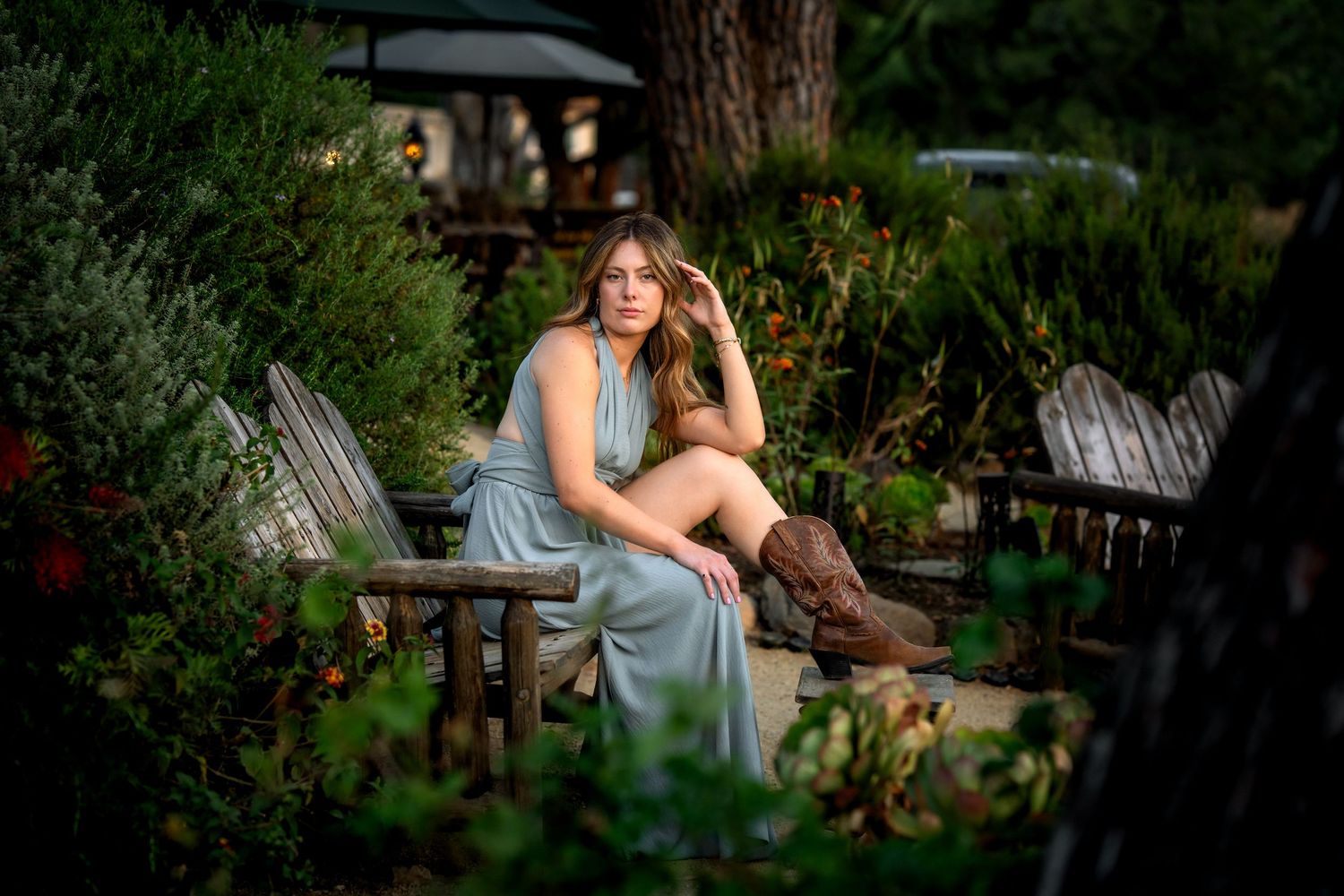 Young woman in light blue dress and cowboy boots sits on rustic wooden bench surrounded by lush garden greenery.