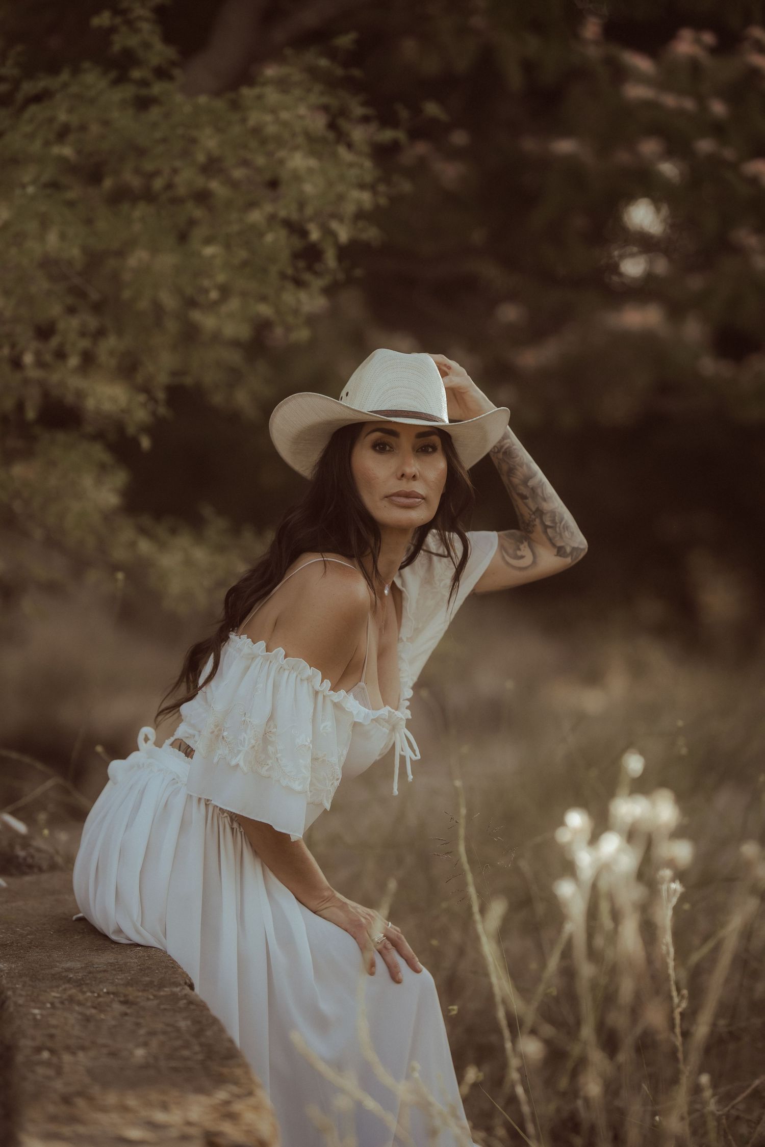 Woman in white off-shoulder dress and cowboy hat poses outdoors in golden light with tattoos visible.