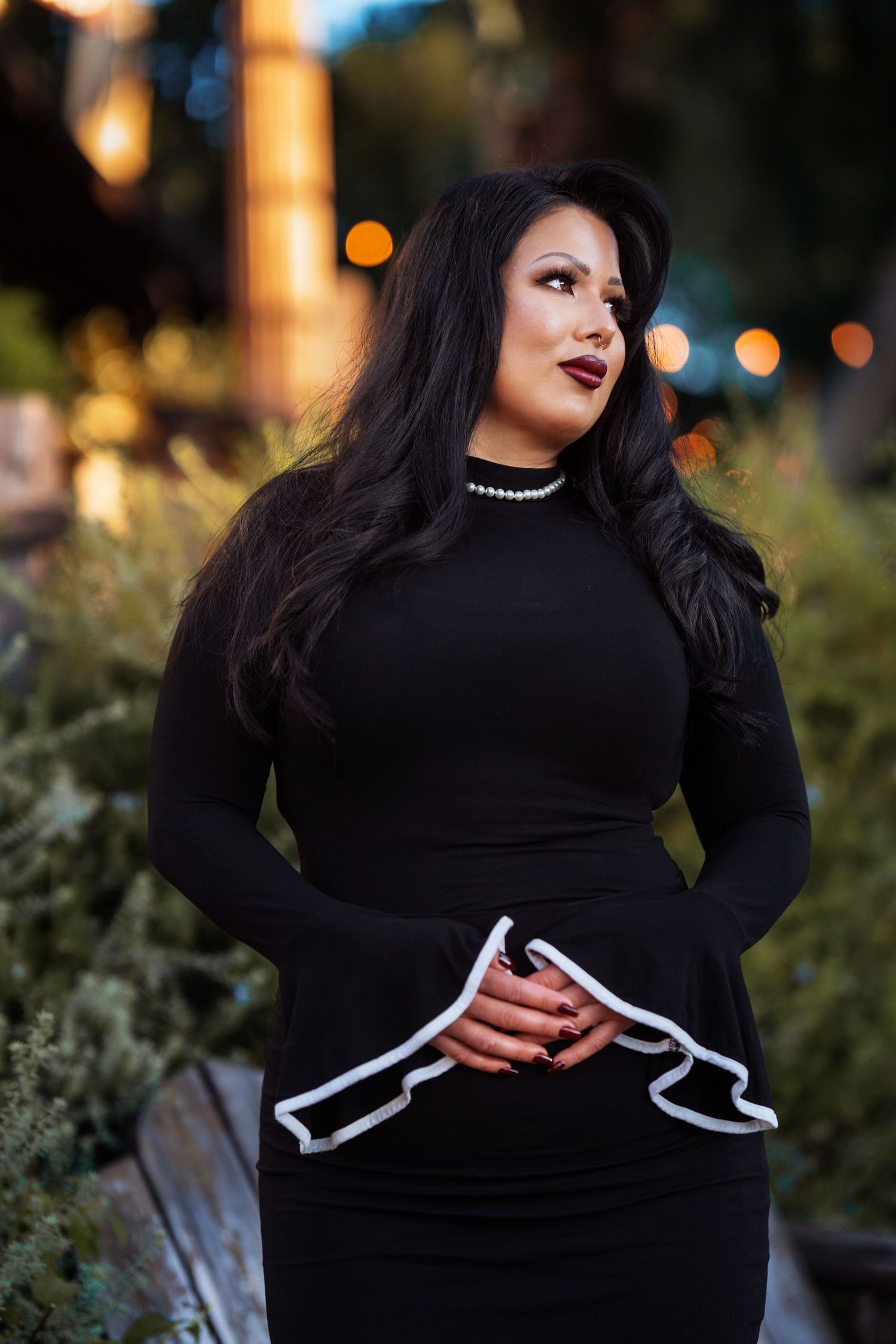 Woman in black long-sleeve dress with gun print detail, pearl necklace, long dark hair, posing outdoors at dusk.