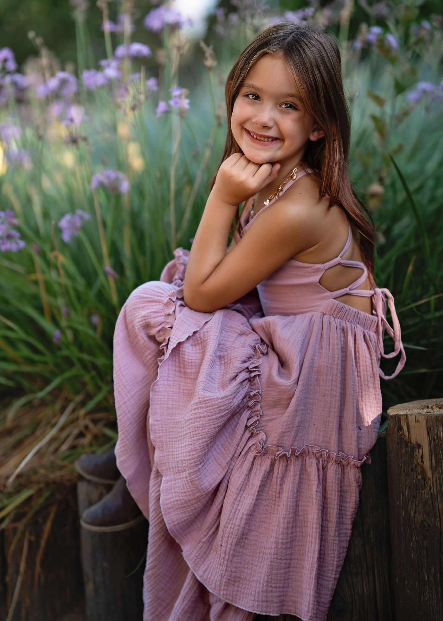 Young girl in purple ruffled dress poses playfully near blooming irises in garden.