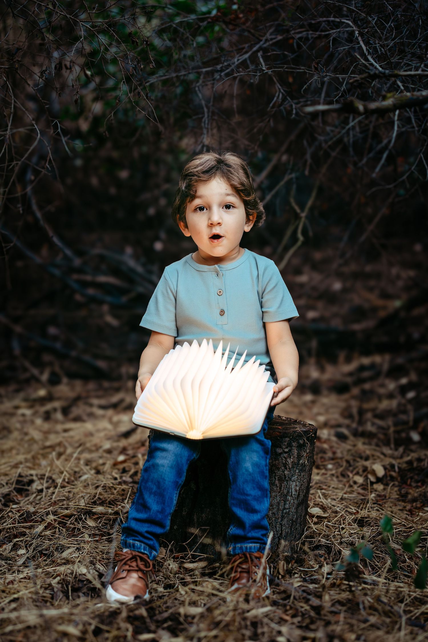 Child sitting on log in woods holding glowing book at dusk.